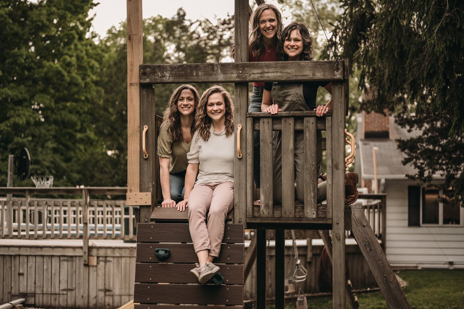 Several people sit and smile on a wooden playground structure in a backyard.