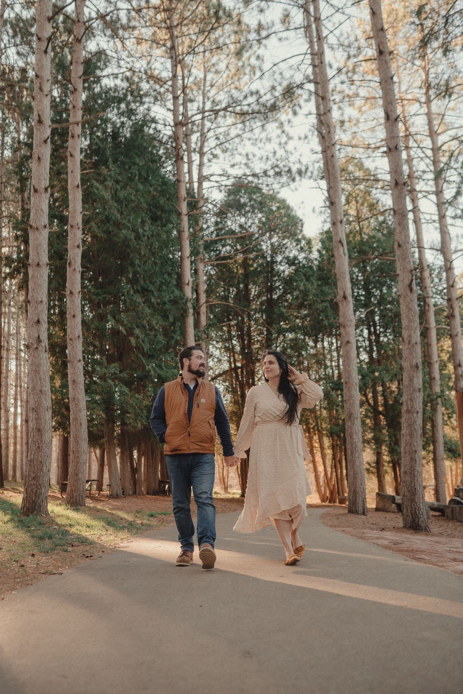 A couple walks hand in hand down a winding forest path surrounded by tall pine trees.