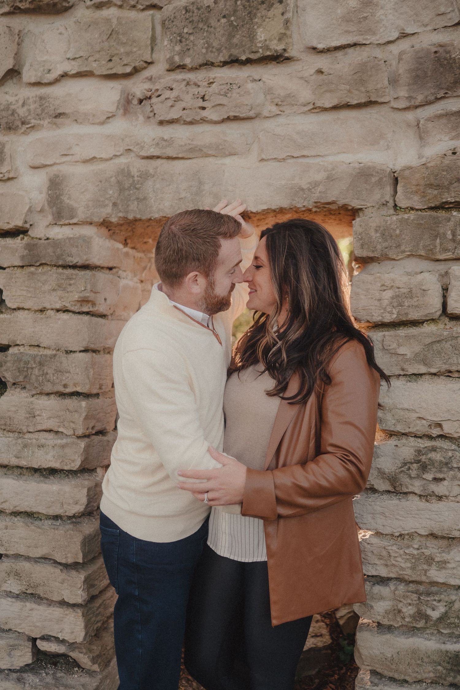 A couple shares an intimate moment against a rustic stone wall backdrop in soft lighting.