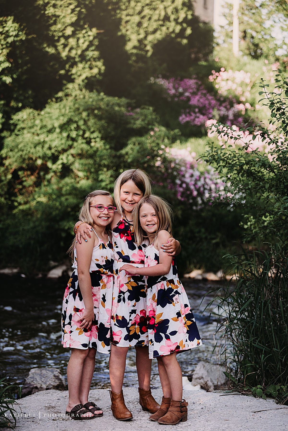 Three people in matching floral dresses pose together on a stone path by a creek with blooming flowers in background.