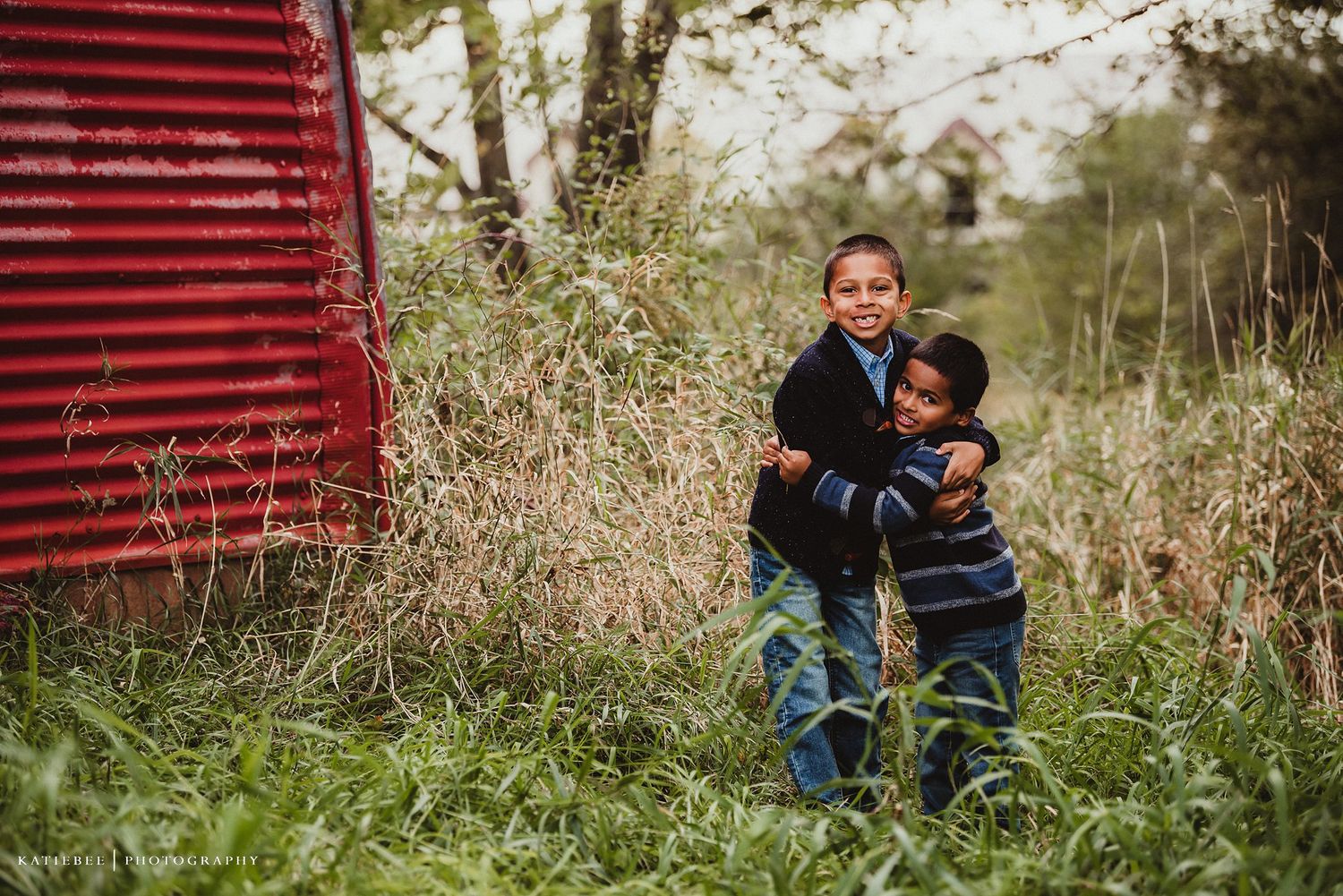 Two people sharing a warm embrace in tall grass beside red barn.