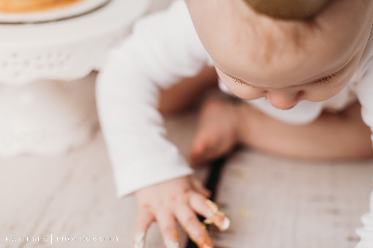 Baby looking down at cake on ornate white cake stand with wreaths on wall.