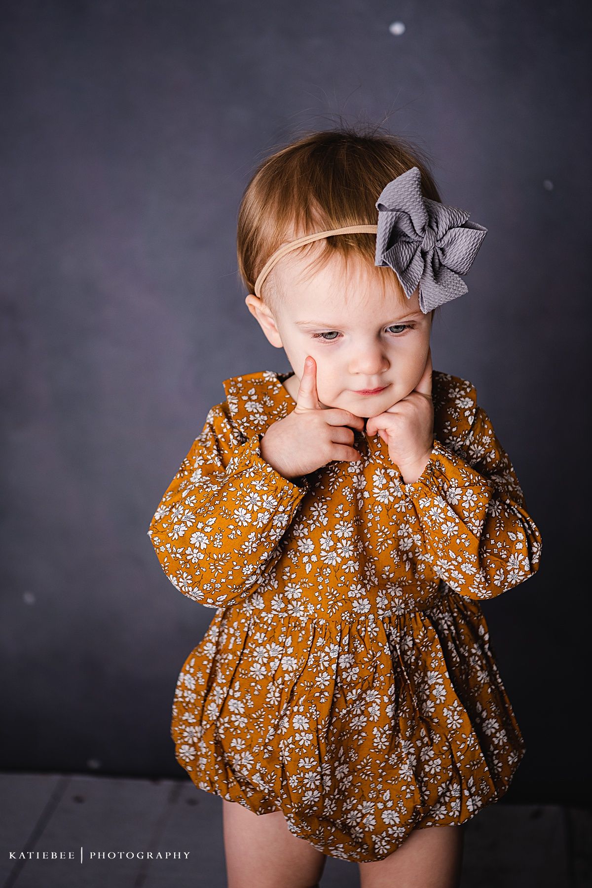 A young child in a floral mustard dress and grey bow makes a playful expression.