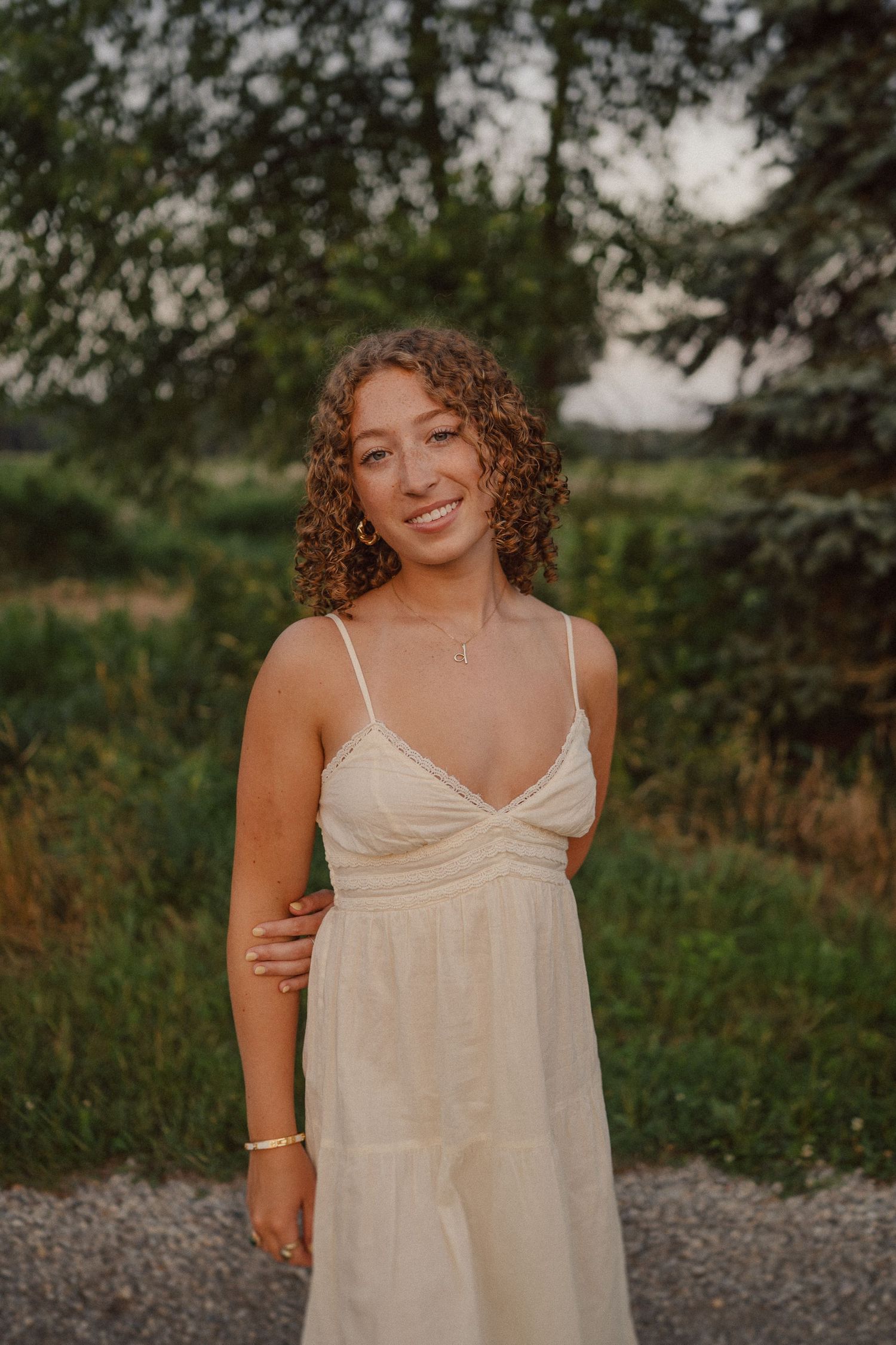A warm sunset portrait of someone wearing a flowing white summer dress standing on a gravel path surrounded by greenery.