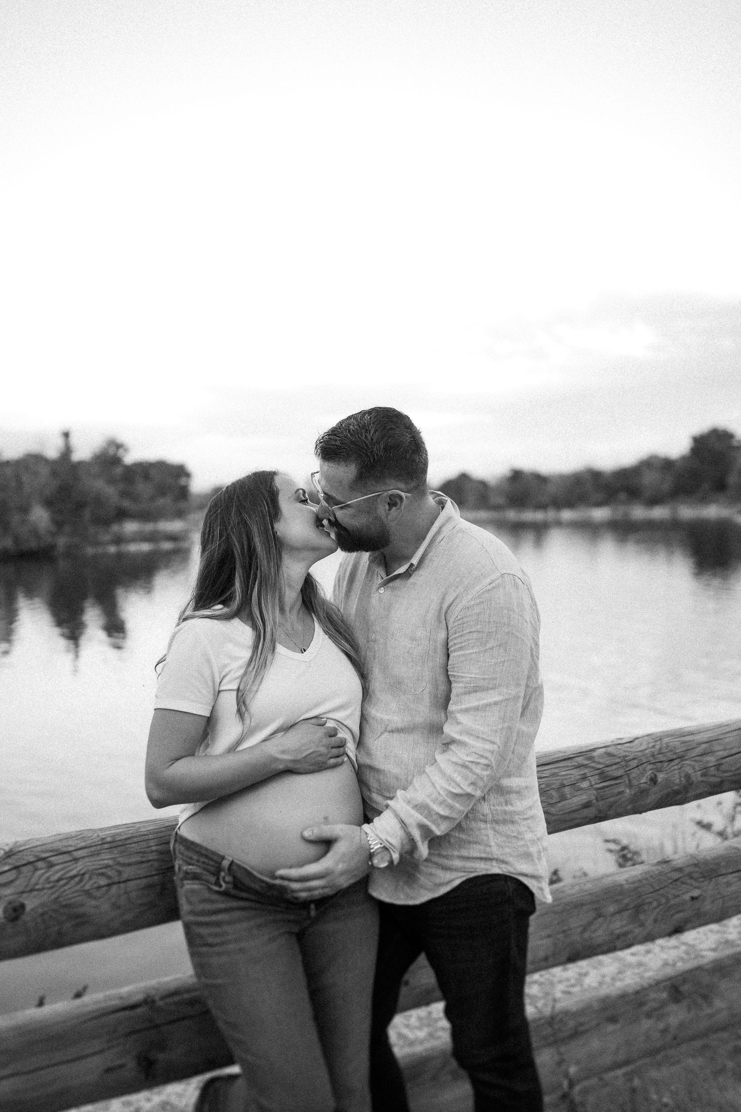 A couple shares a tender moment during their maternity photoshoot by a serene lake with a wooden fence in black and white.