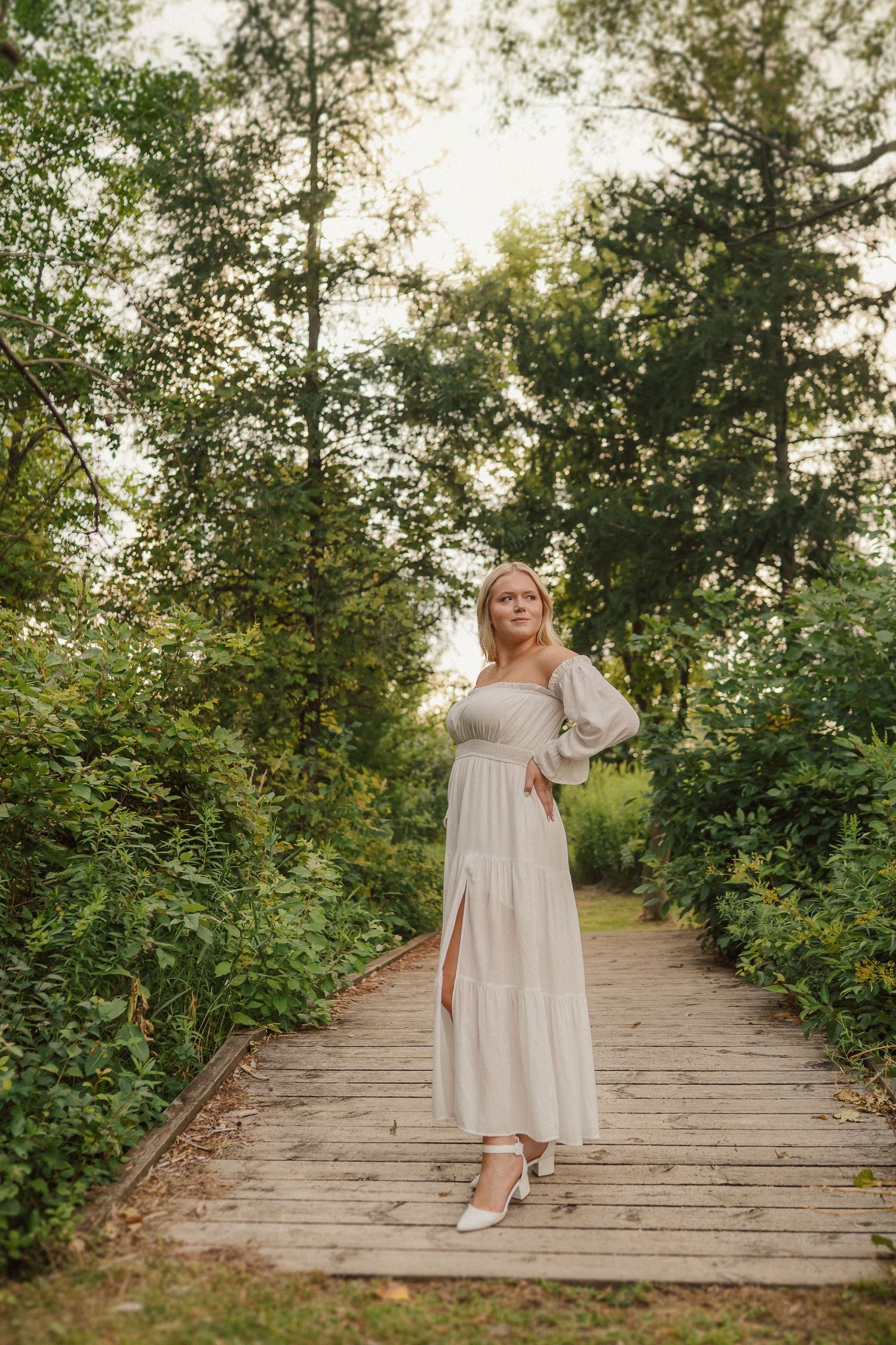 Person in vintage-style white dress poses on wooden boardwalk surrounded by lush greenery.