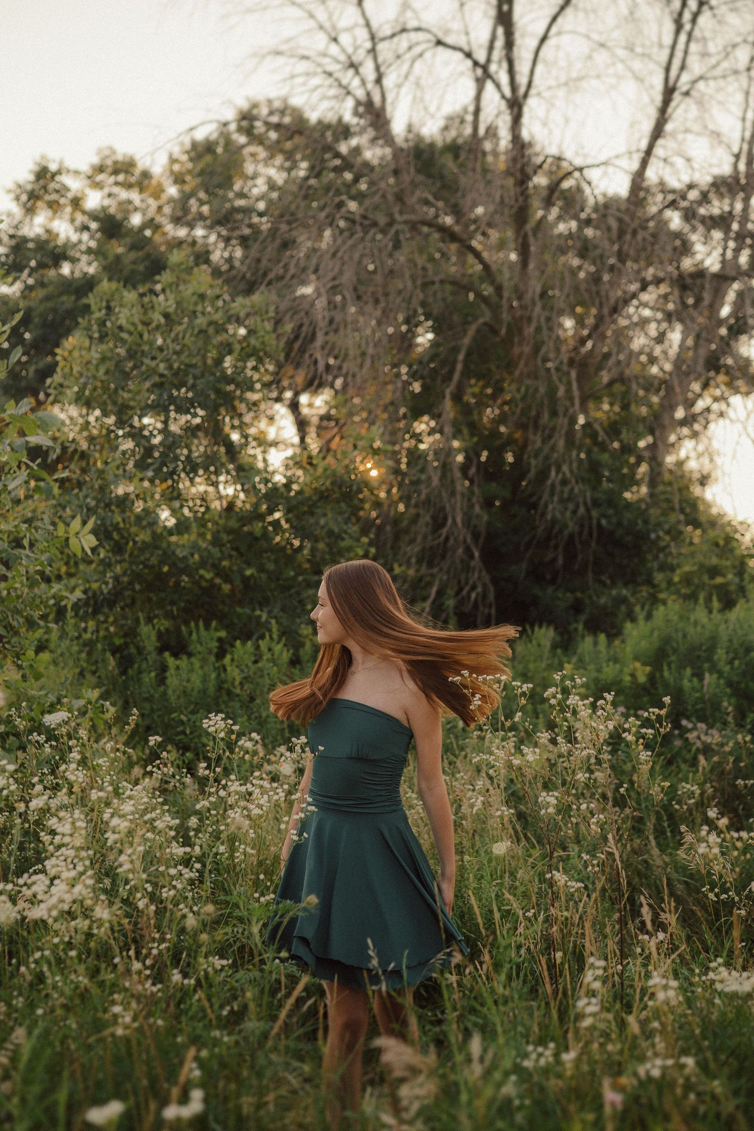A green dress flows through a dreamy wildflower field at sunset with tall grass and trees in background.