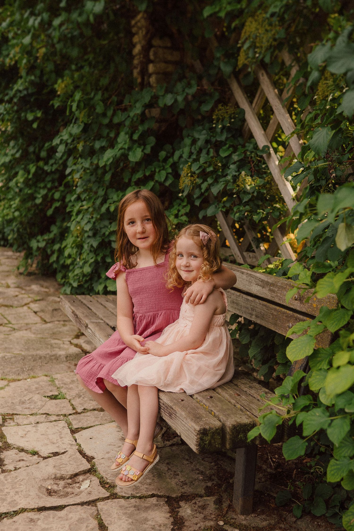 Two figures in pink dresses share a sweet moment together on a rustic wooden bench surrounded by lush greenery.