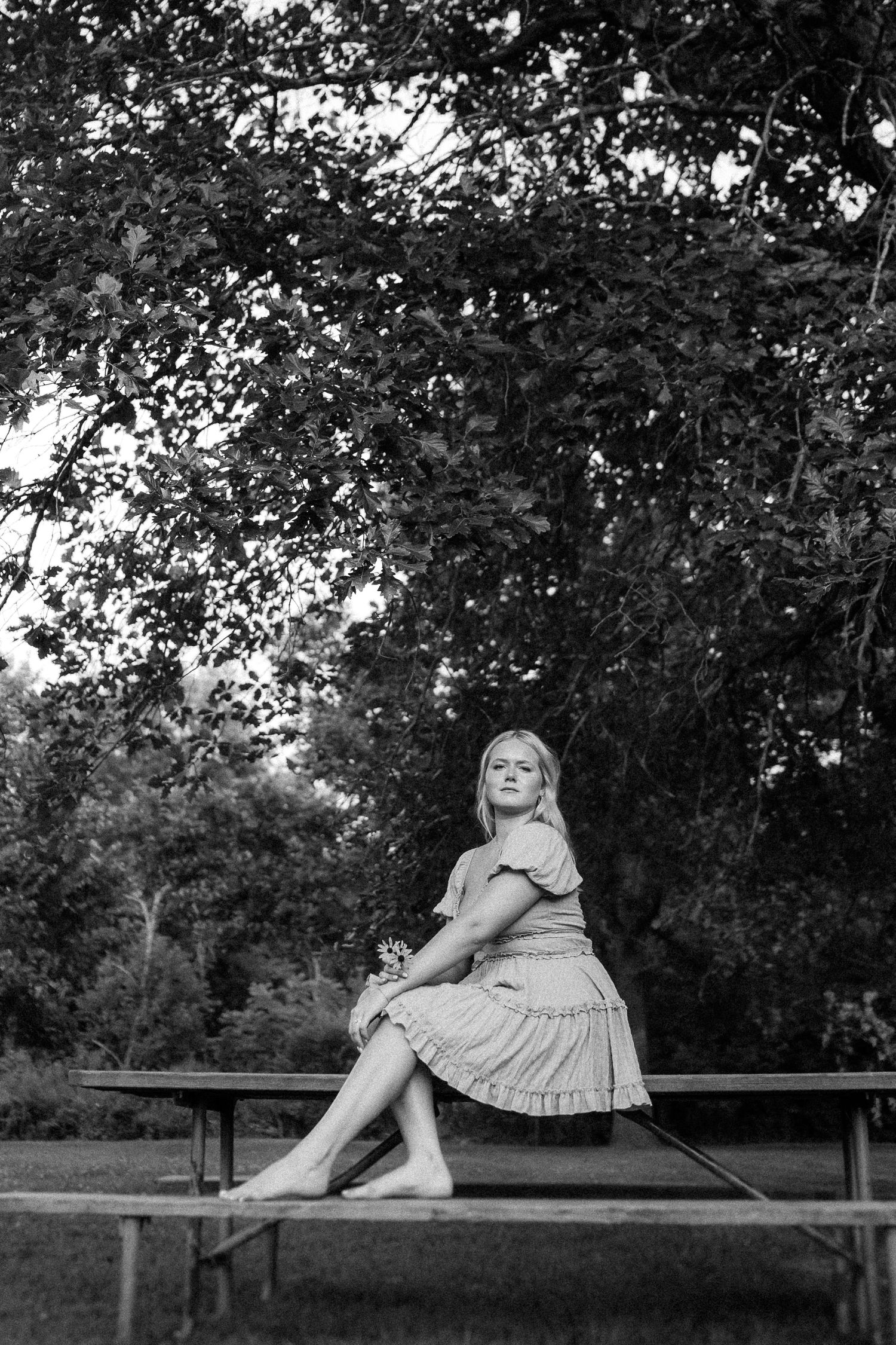 A black and white portrait of someone sitting on a picnic table beneath tree branches in a park setting.