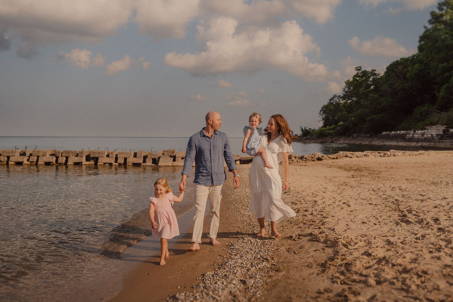 A family takes an evening walk along the shoreline with wooden posts visible in the water.