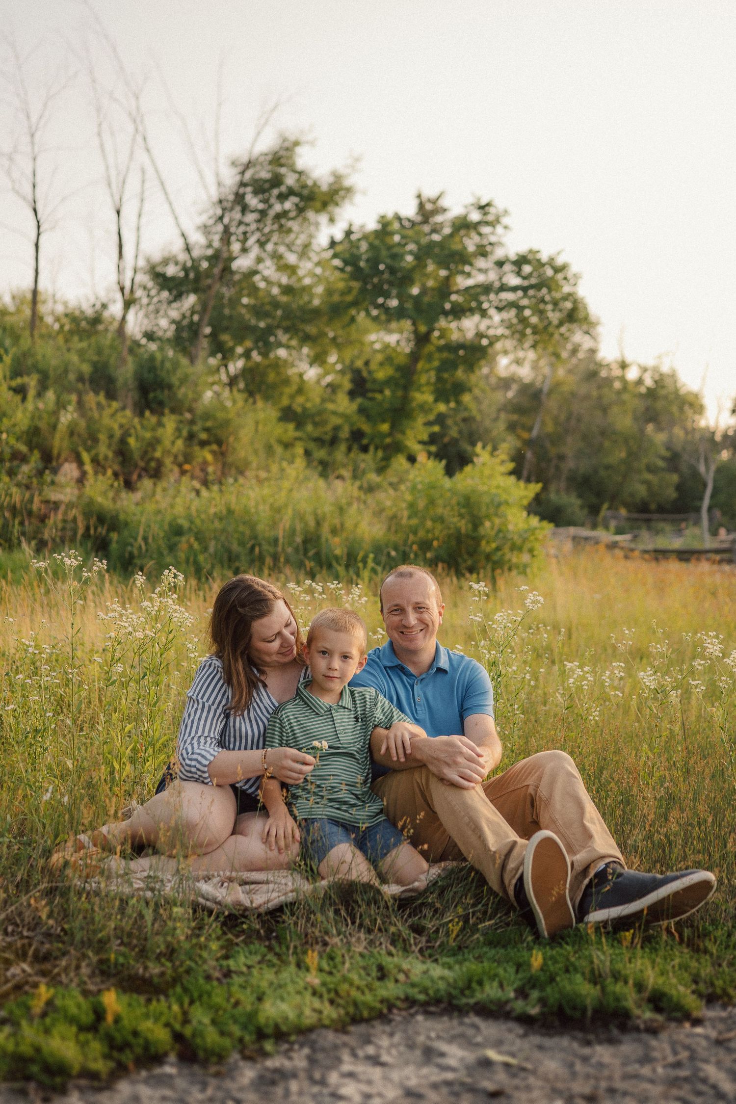 Family relaxing together in a sun-lit meadow with tall grass and trees in the background.