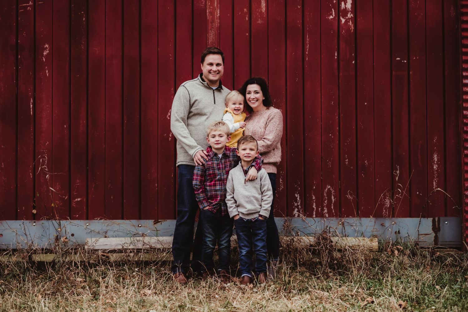 A family of five poses for a portrait against a rustic red barn wall.