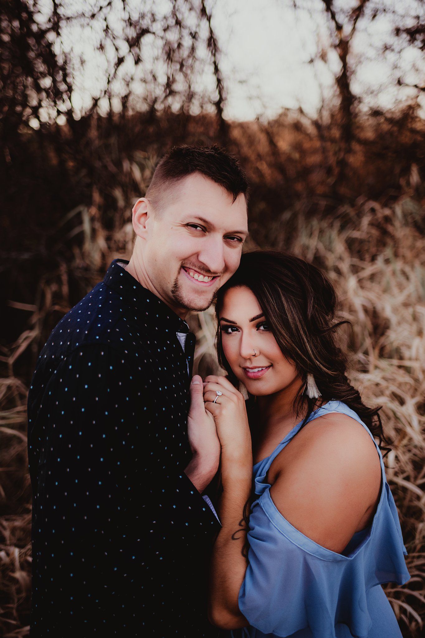 Stylish couple in blue dress and polka dot shirt pose together in autumn field at sunset.