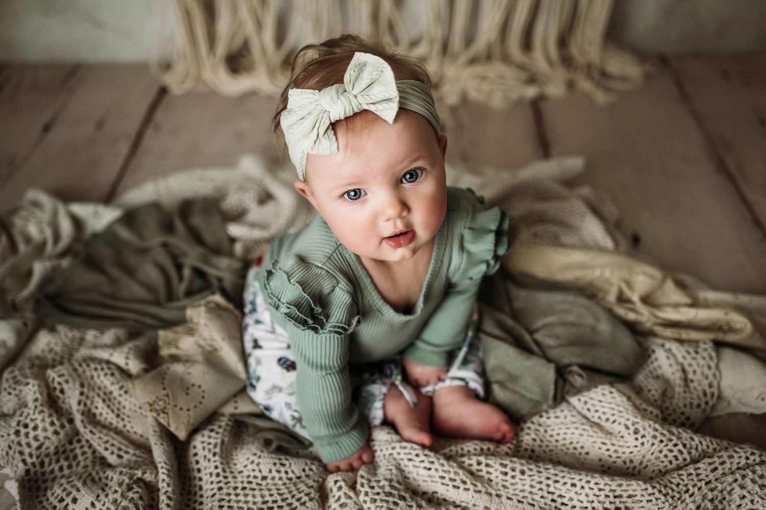 Baby in sage green outfit with white bow sits on neutral toned blankets with macrame wall decor.