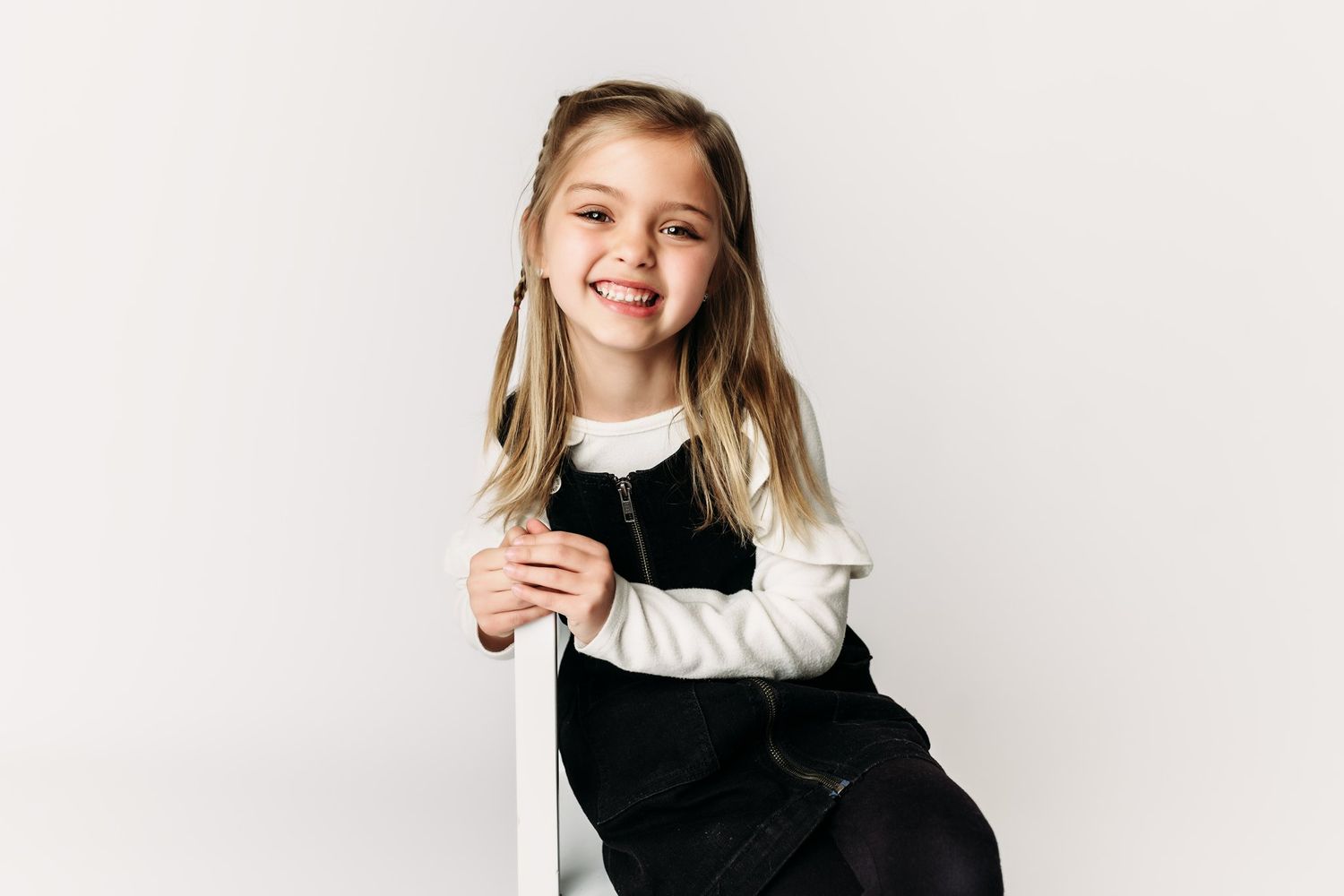 A child sits casually on a white stool in a black and white ensemble.