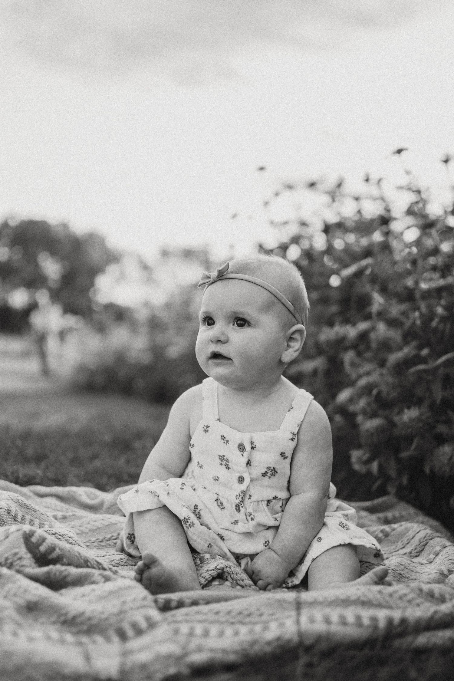Black and white photo of baby in patterned outfit sitting on blanket outdoors.