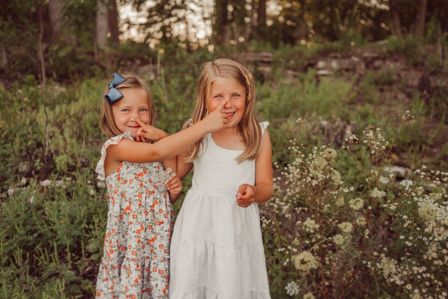 Two children in summer dresses pose together among wildflowers at sunset.
