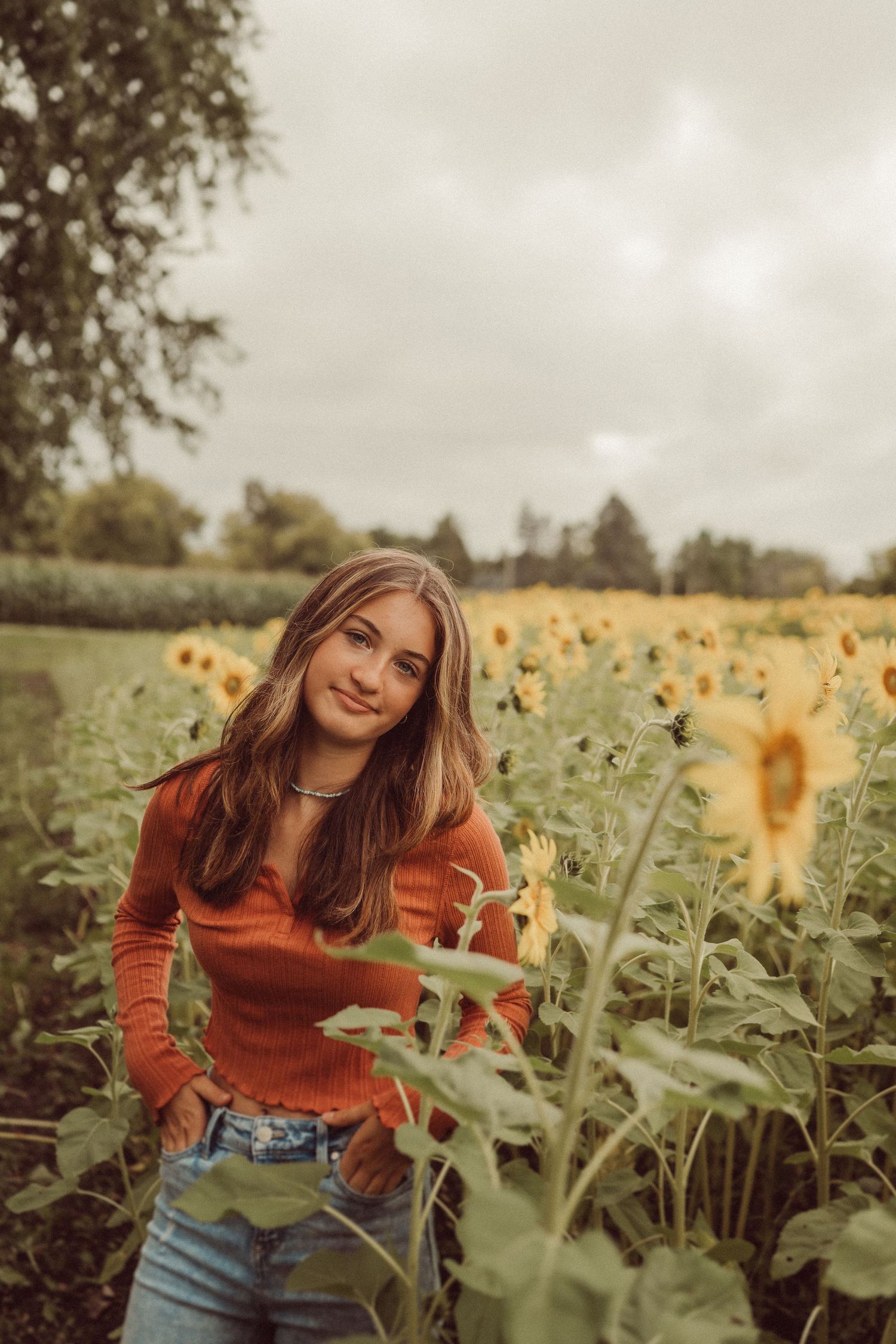 A person in a rust-colored sweater poses casually in a field of blooming sunflowers.