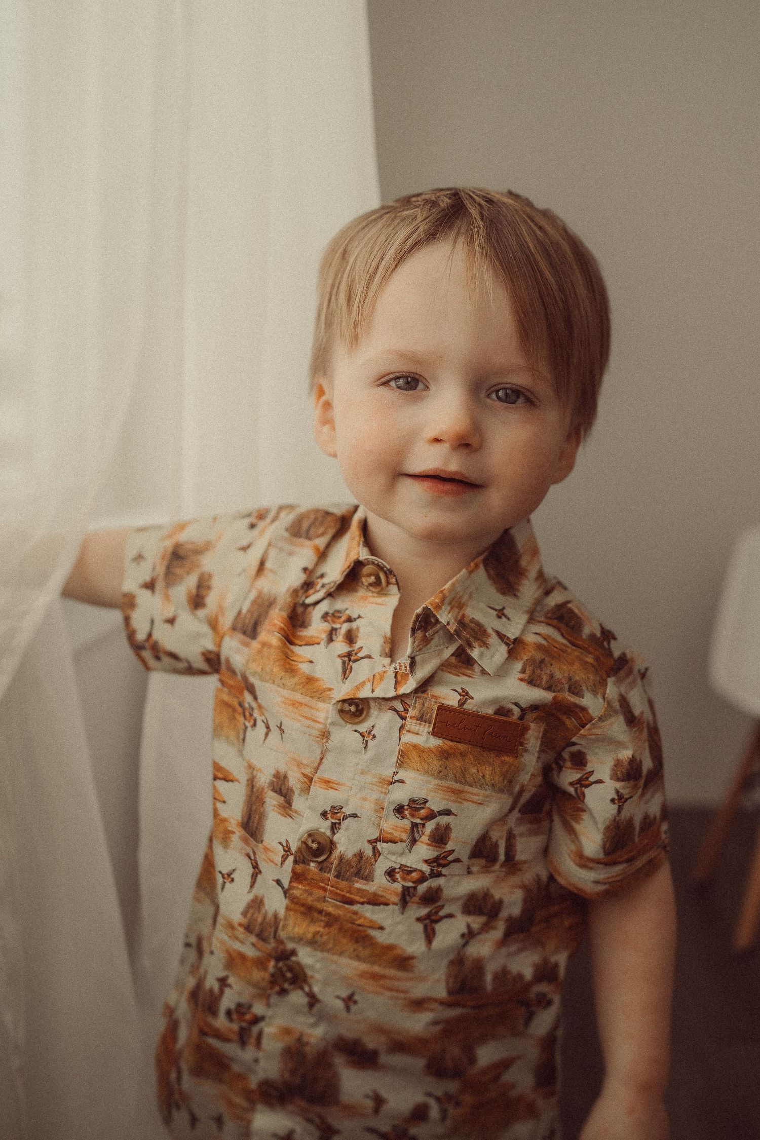 Portrait of toddler wearing brown floral button-up shirt against white backdrop.