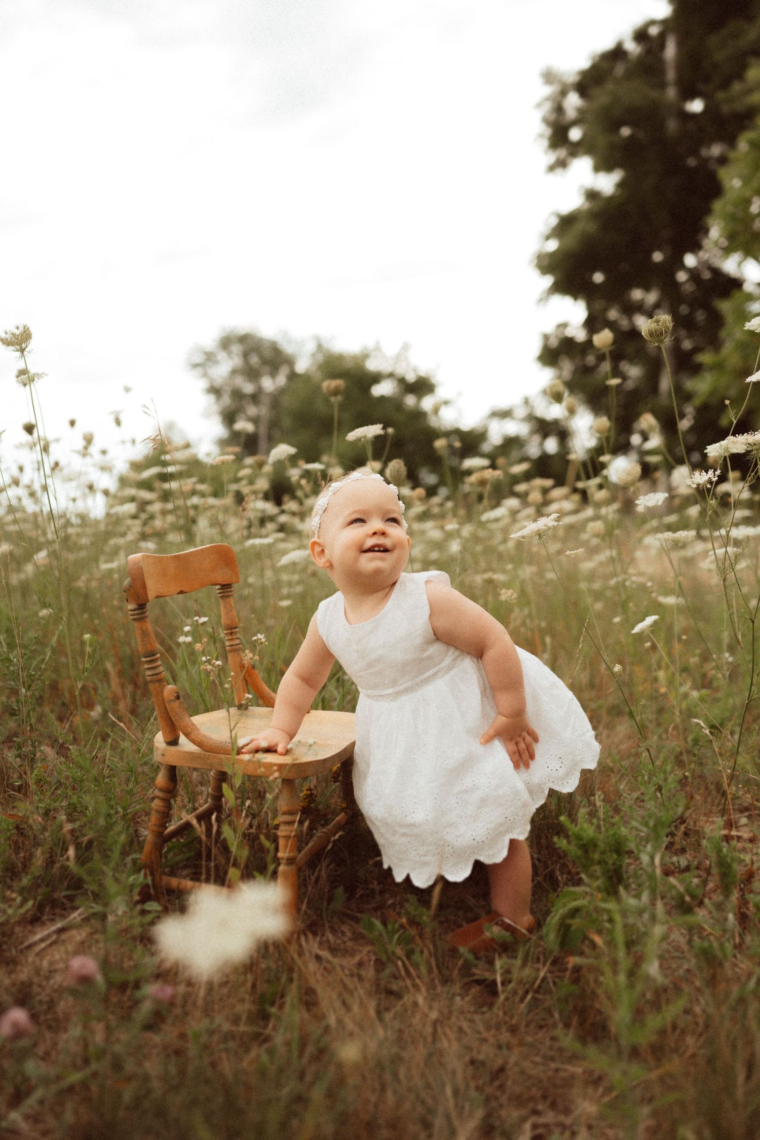 Baby in white dress sits on wooden chair in field of wildflowers.