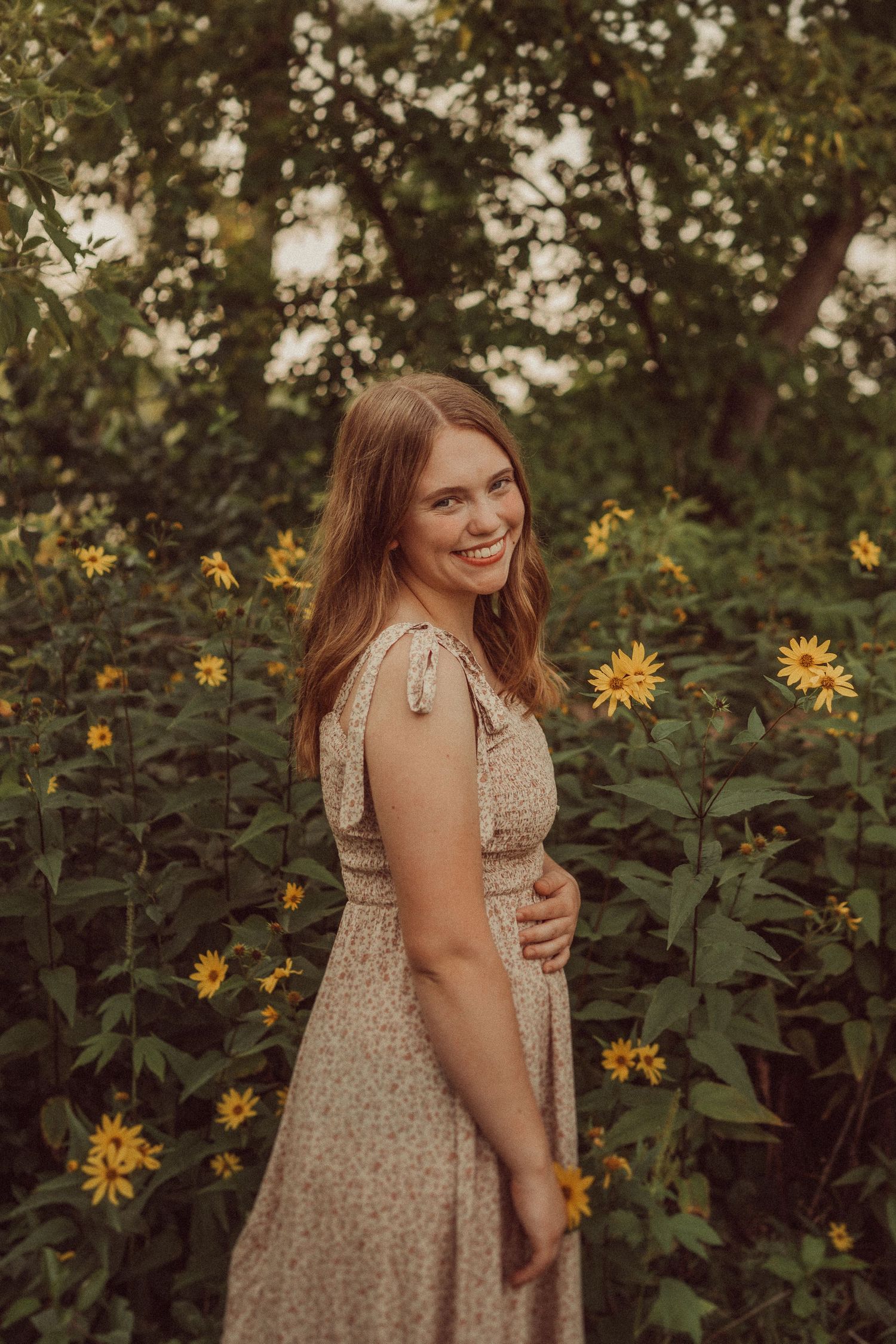A person in a floral dress poses in a garden with blooming sunflowers on a warm summer evening.