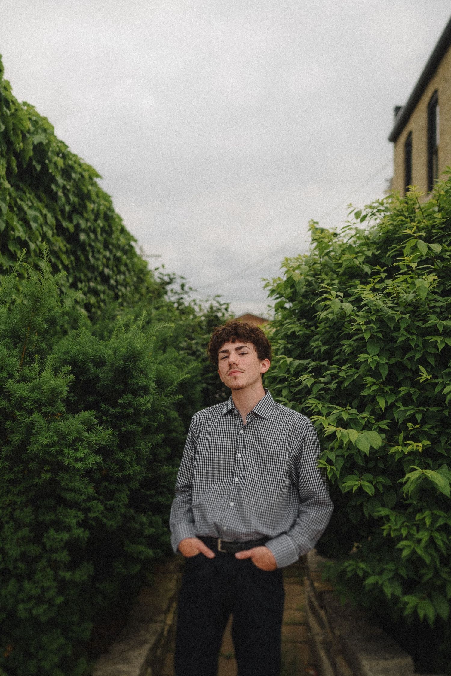 A person in a checkered shirt poses between green hedgerows on an overcast day.