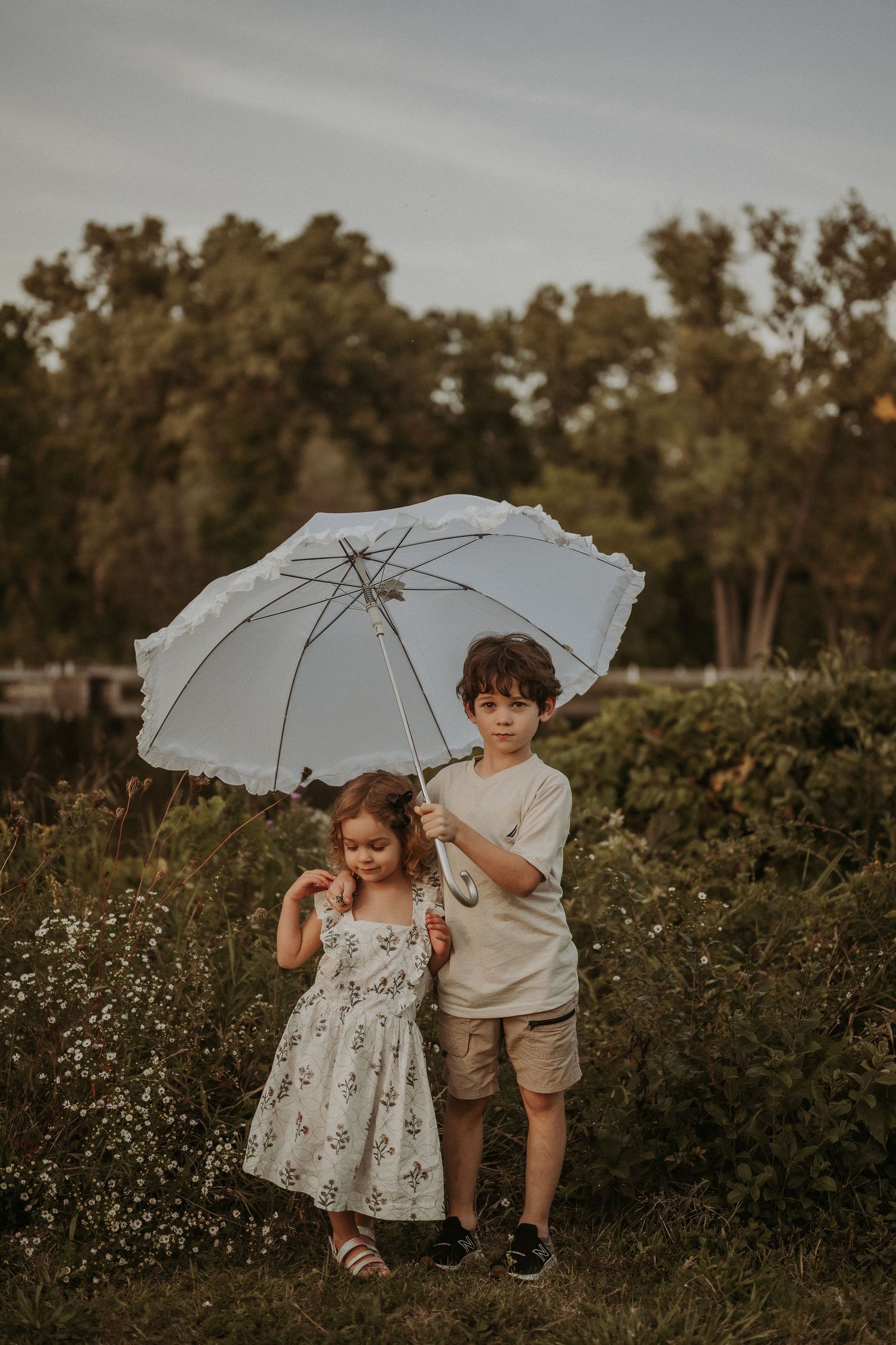 Two children pose with a white umbrella in a meadow during sunset.