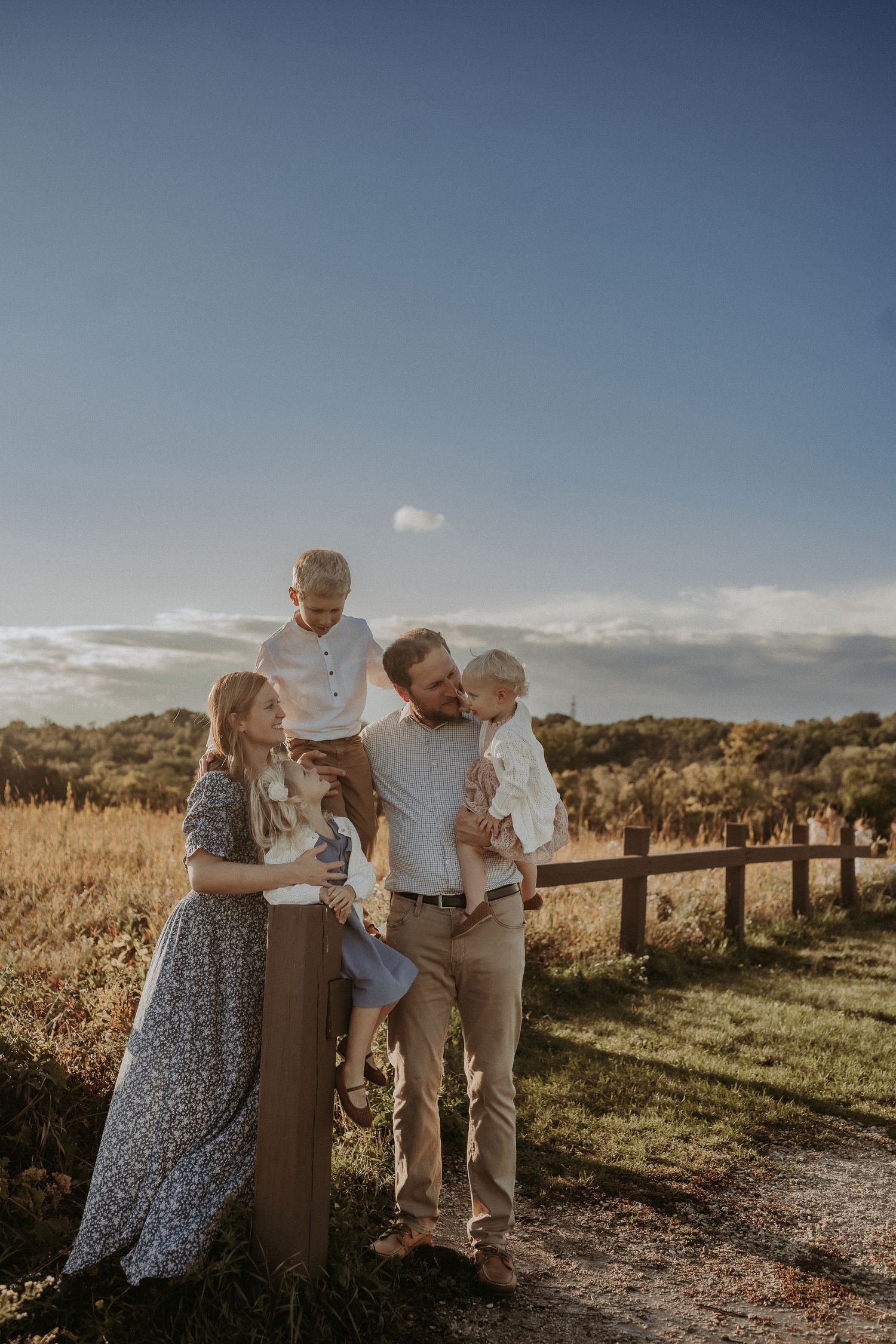 Family dressed in coordinating outfits posing together by a wooden fence during golden hour.