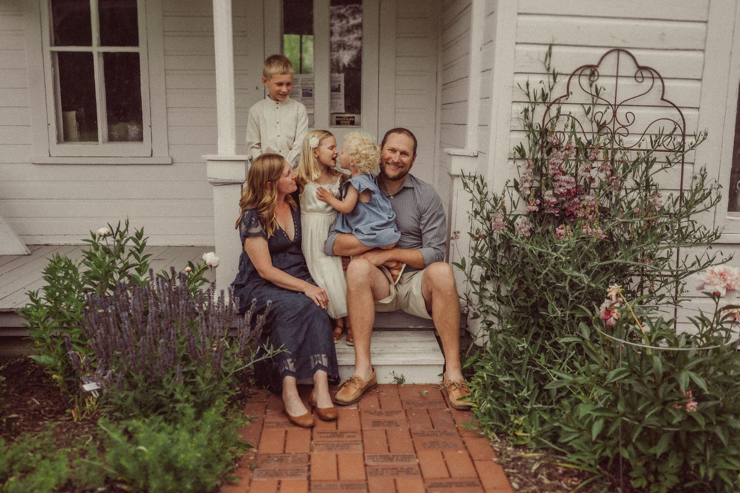 Casual family moment captured on brick steps of a white farmhouse with blooming garden.