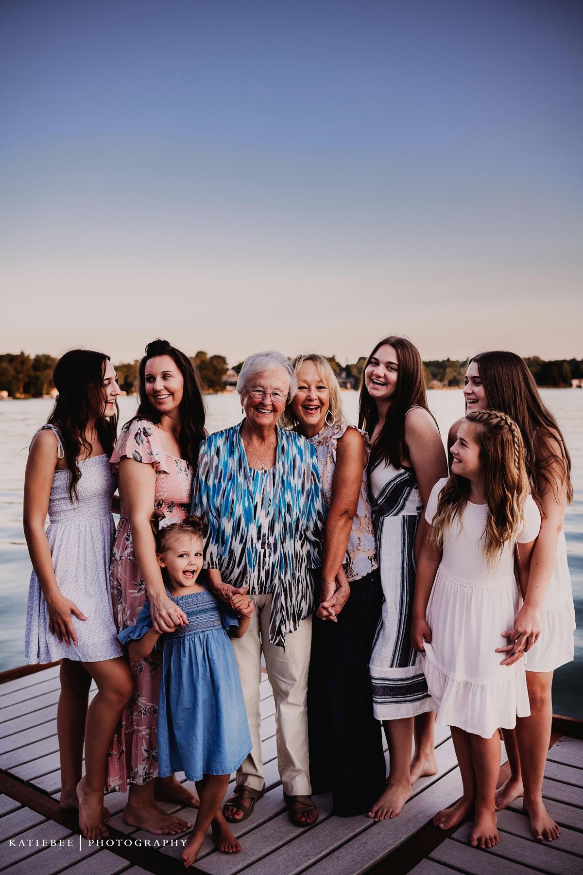 Family group in summer dresses poses on wooden dock at sunset with water and trees in background.
