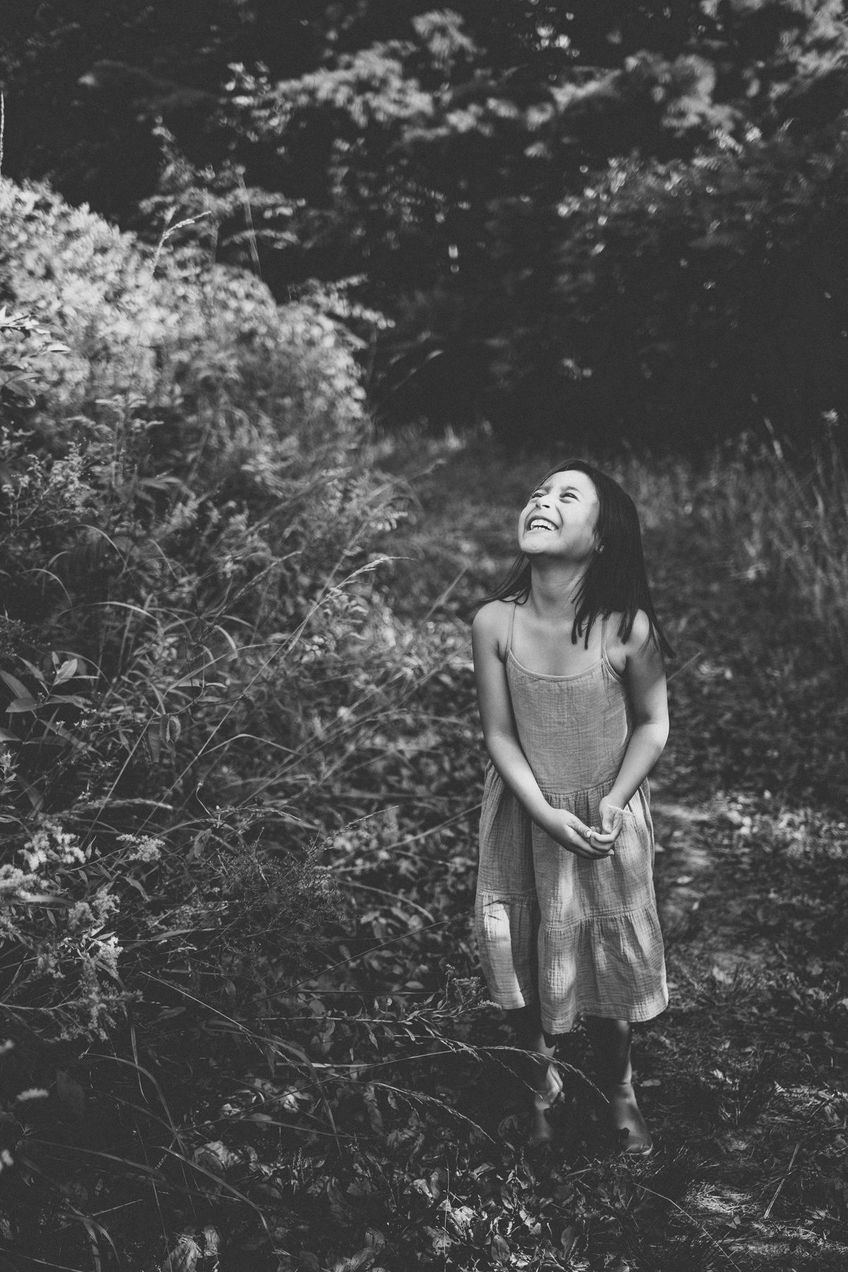 Black and white portrait of someone laughing joyfully in a garden setting wearing a casual dress.