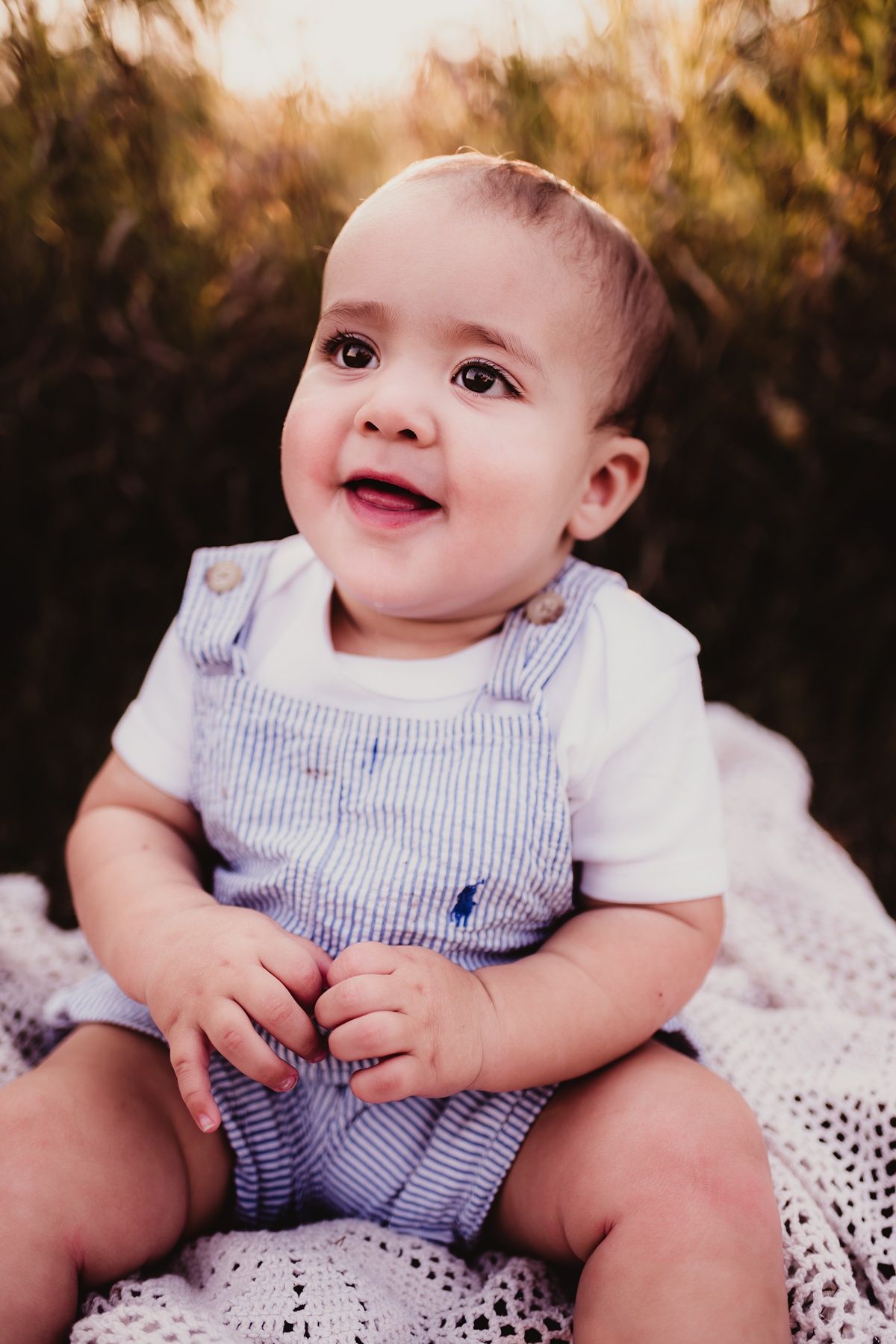 Baby wearing blue and white striped overalls sits on white blanket during golden hour sunset.