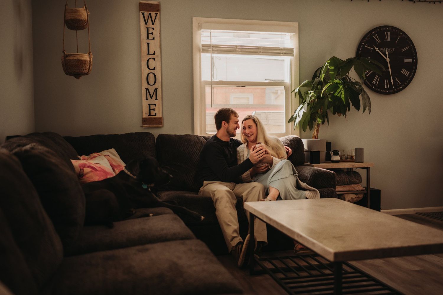 A cozy living room scene with a couple relaxing on a dark couch near a window with natural light.