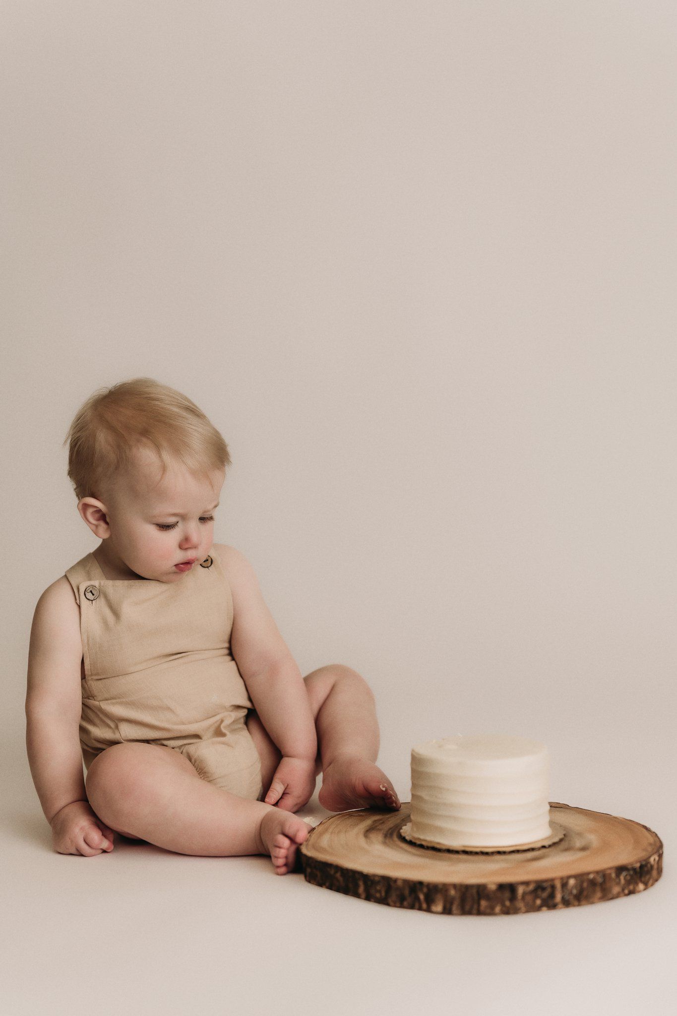 Emotional baby in beige outfit next to minimalist white cake on wood slice against neutral background.