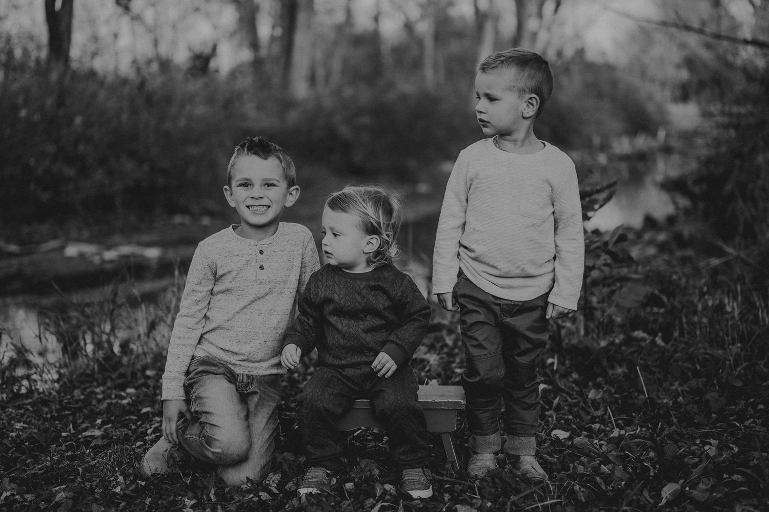 Three children sit together in a black and white outdoor portrait surrounded by nature.