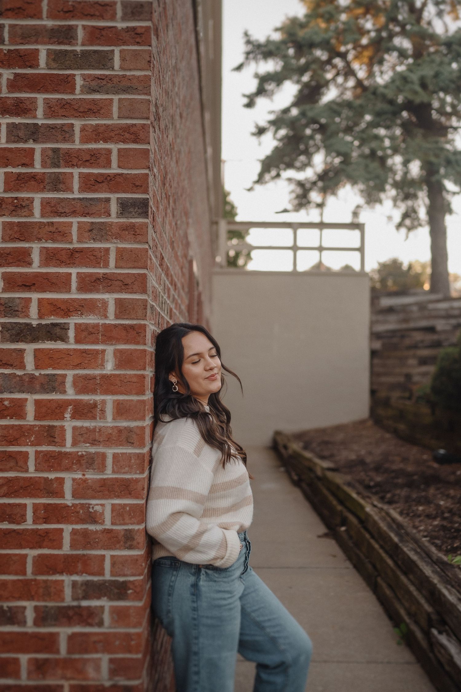 Person in striped sweater and jeans leaning against brick wall in outdoor setting.