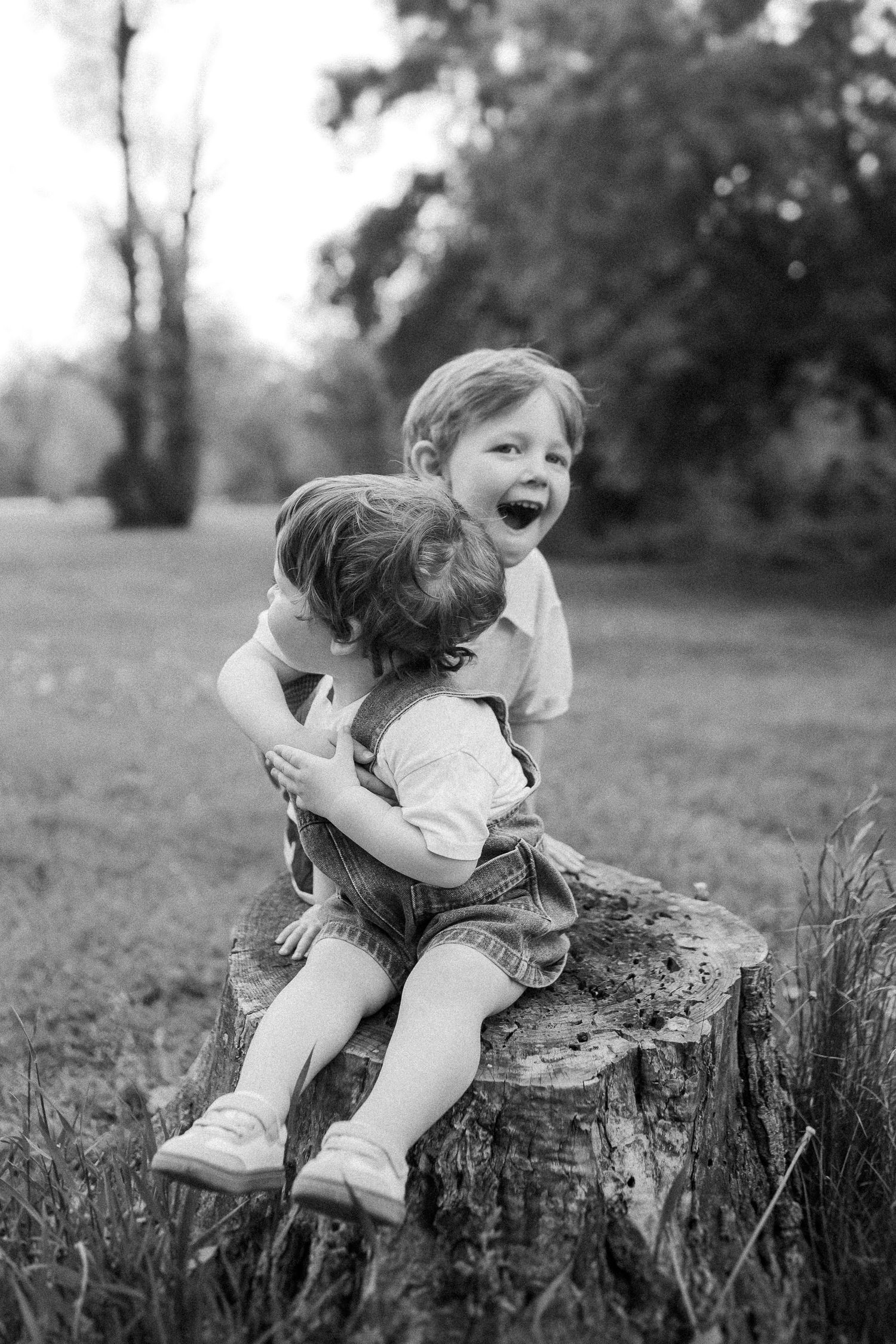 Two children sharing a joyful moment while sitting on a tree stump in a black and white photograph.