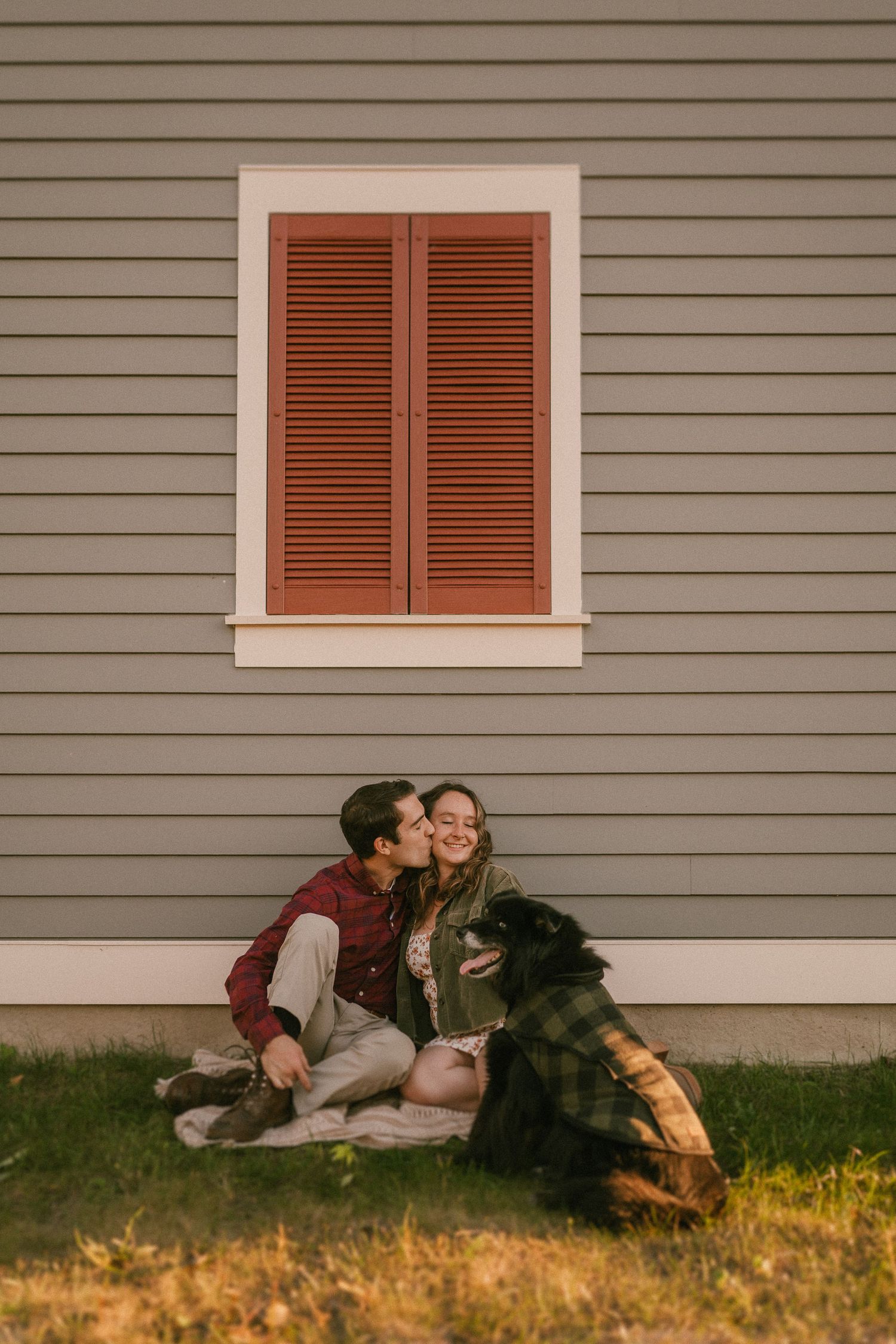 Couple sits together against gray house siding with brown shutter window during golden hour.