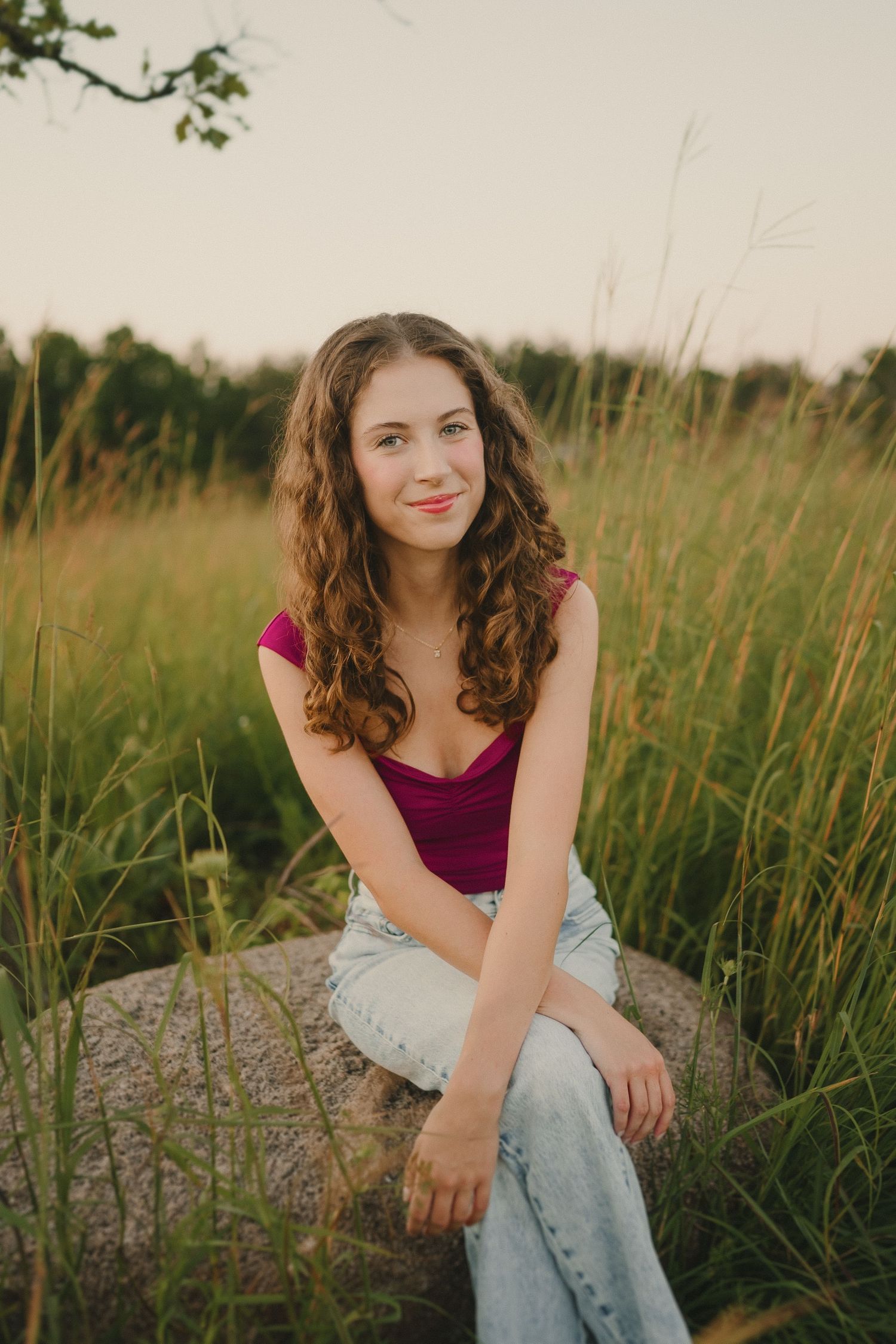 A casual portrait shows someone sitting on a rock amid tall grass wearing a burgundy top and jeans.