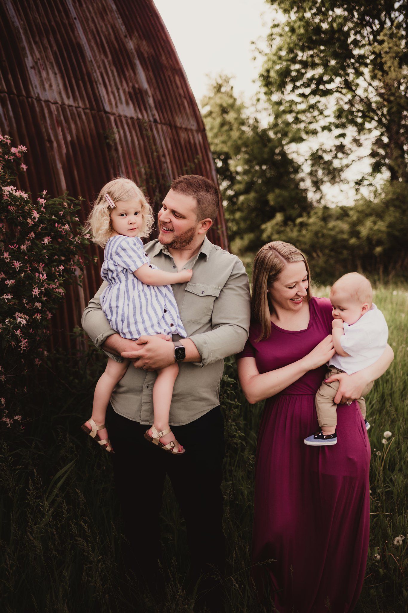 Family portrait in front of a rustic barn with flowering bushes at sunset.