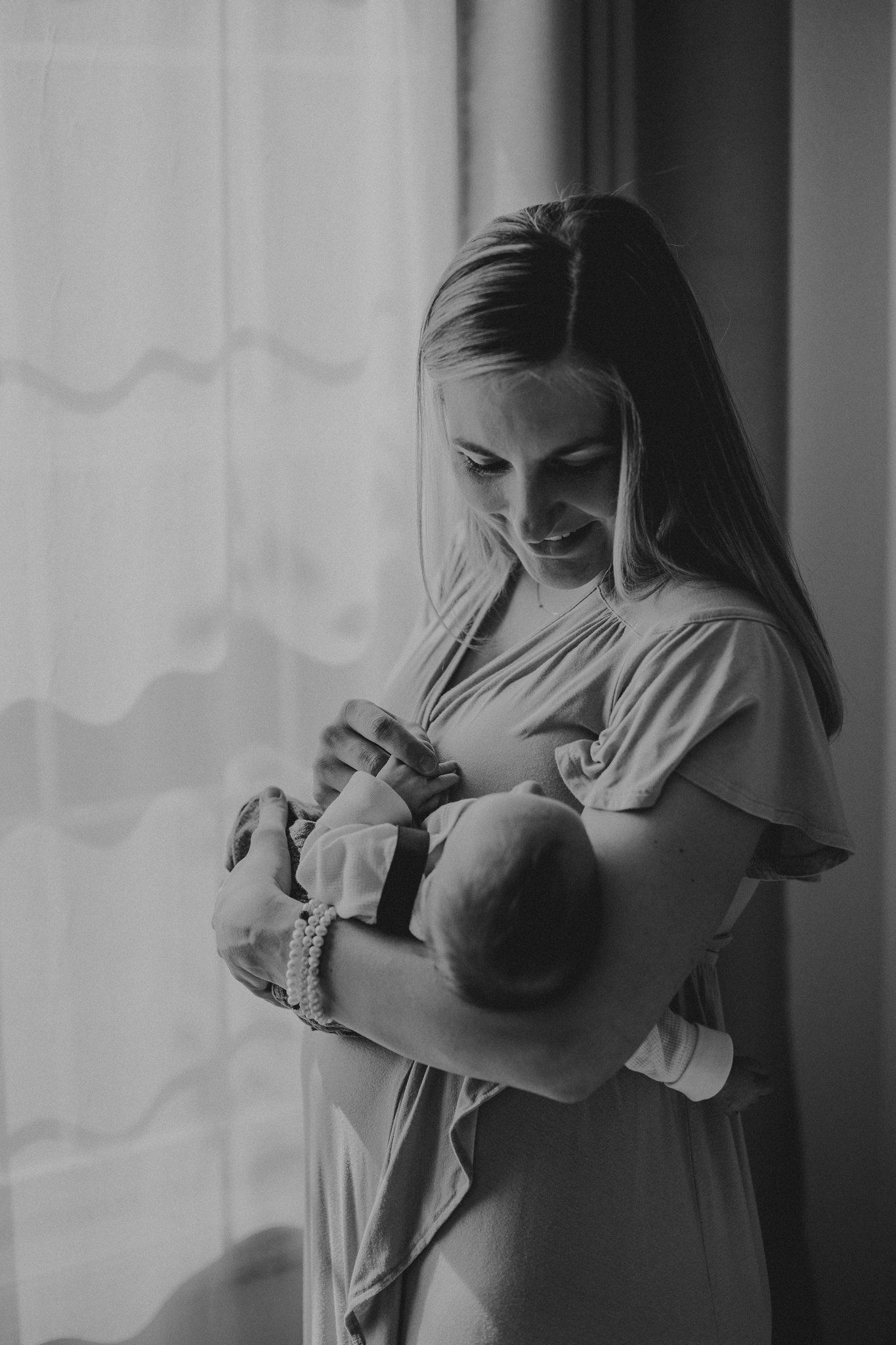 Mother in white dress cradles newborn baby near window with natural light.