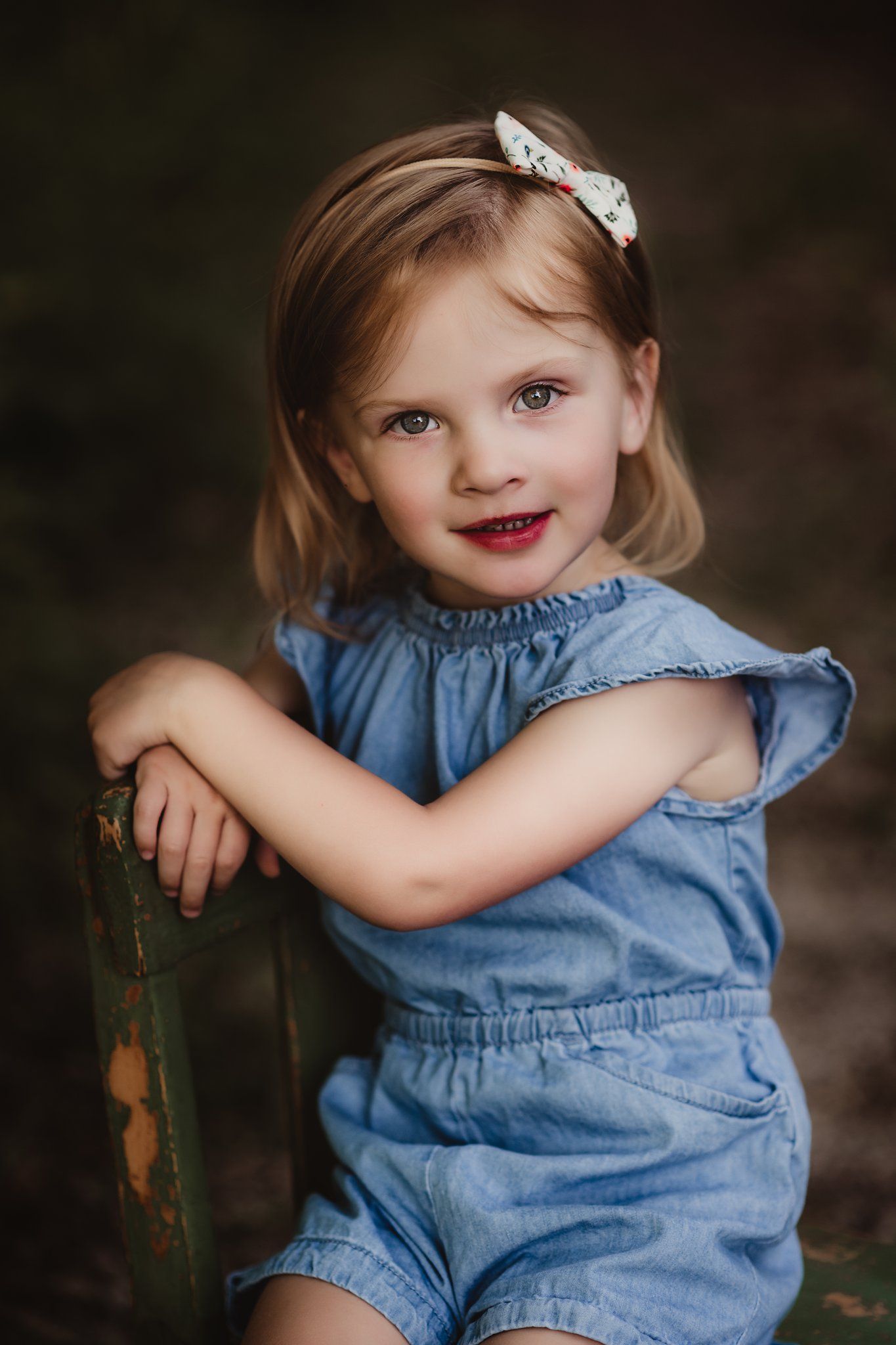 Child in denim romper posing against dark background with a white bow in their hair.