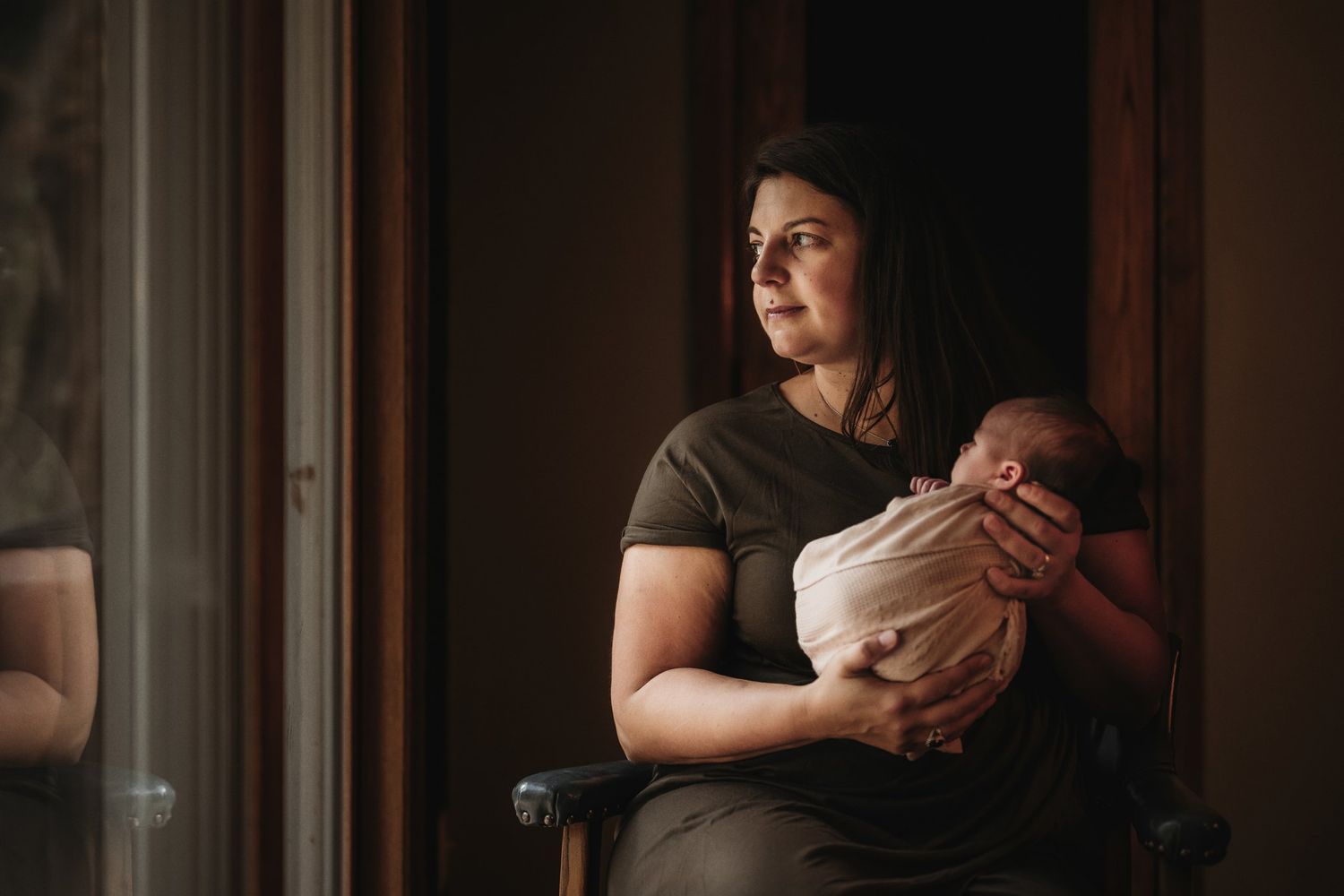 Mother in olive dress holds sleeping newborn by window in moody lighting.