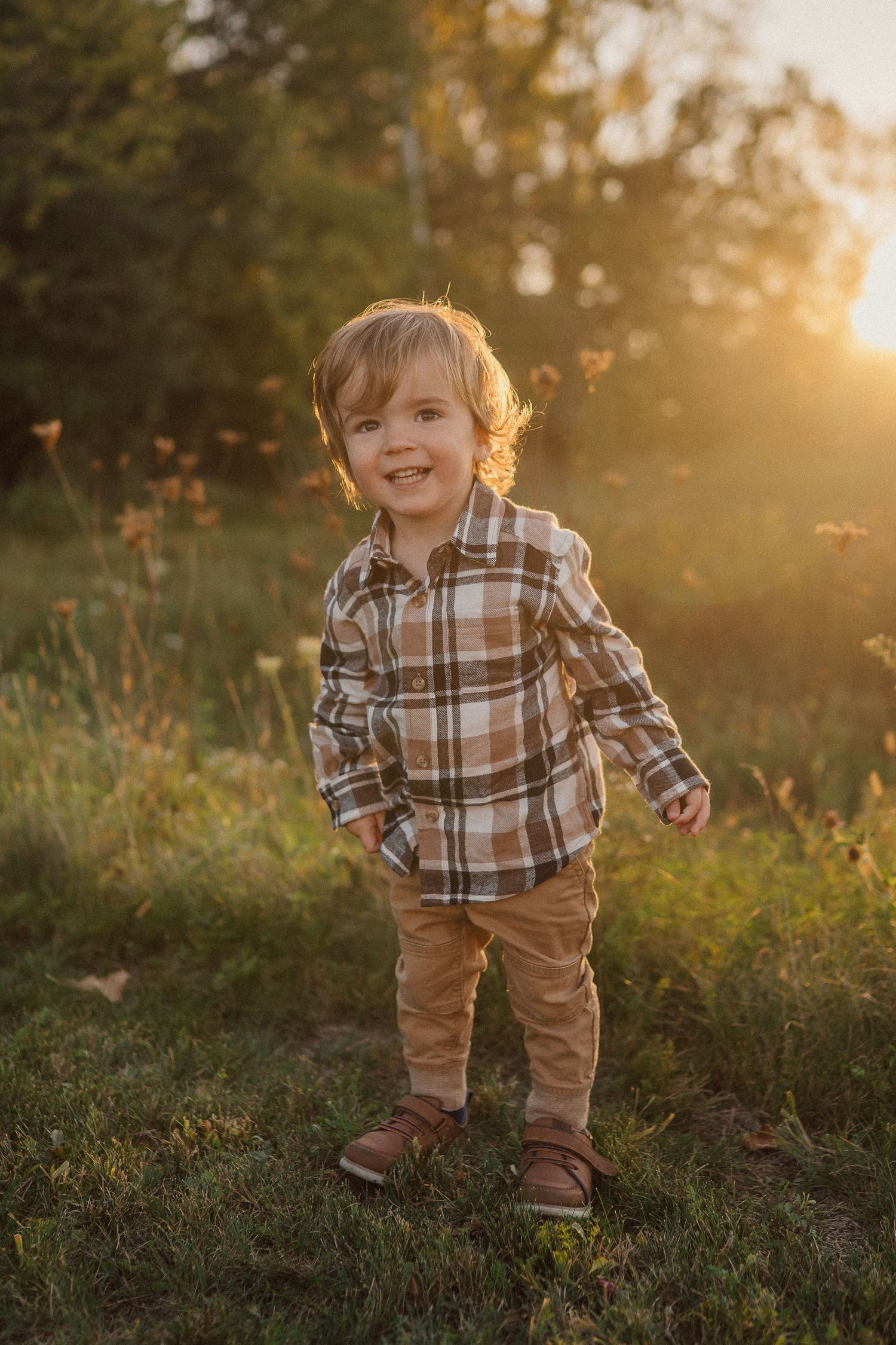 A smiling child in plaid and brown pants plays in a grassy field at sunset.