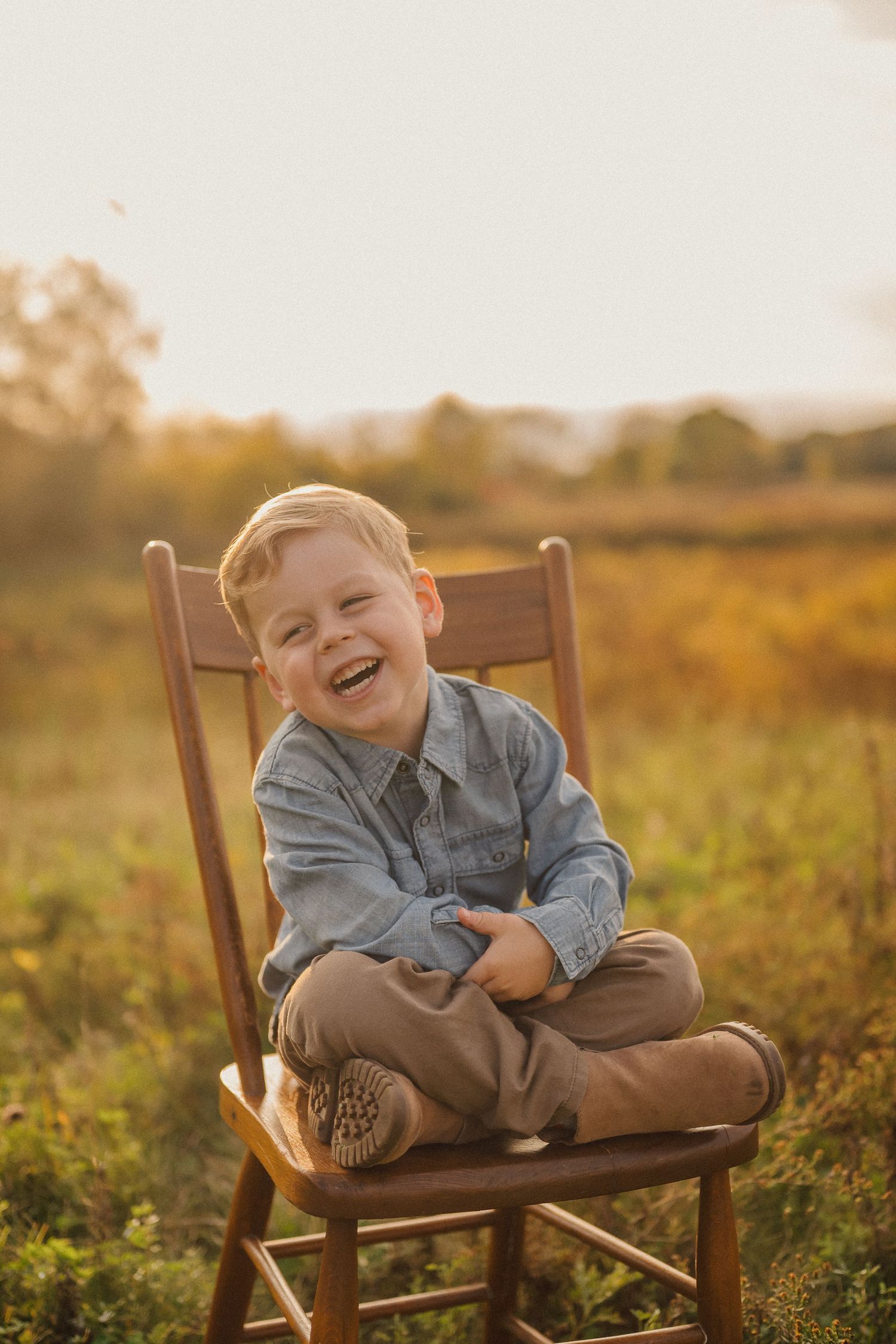 A young child laughs while sitting on a wooden chair in a sunlit autumn field.