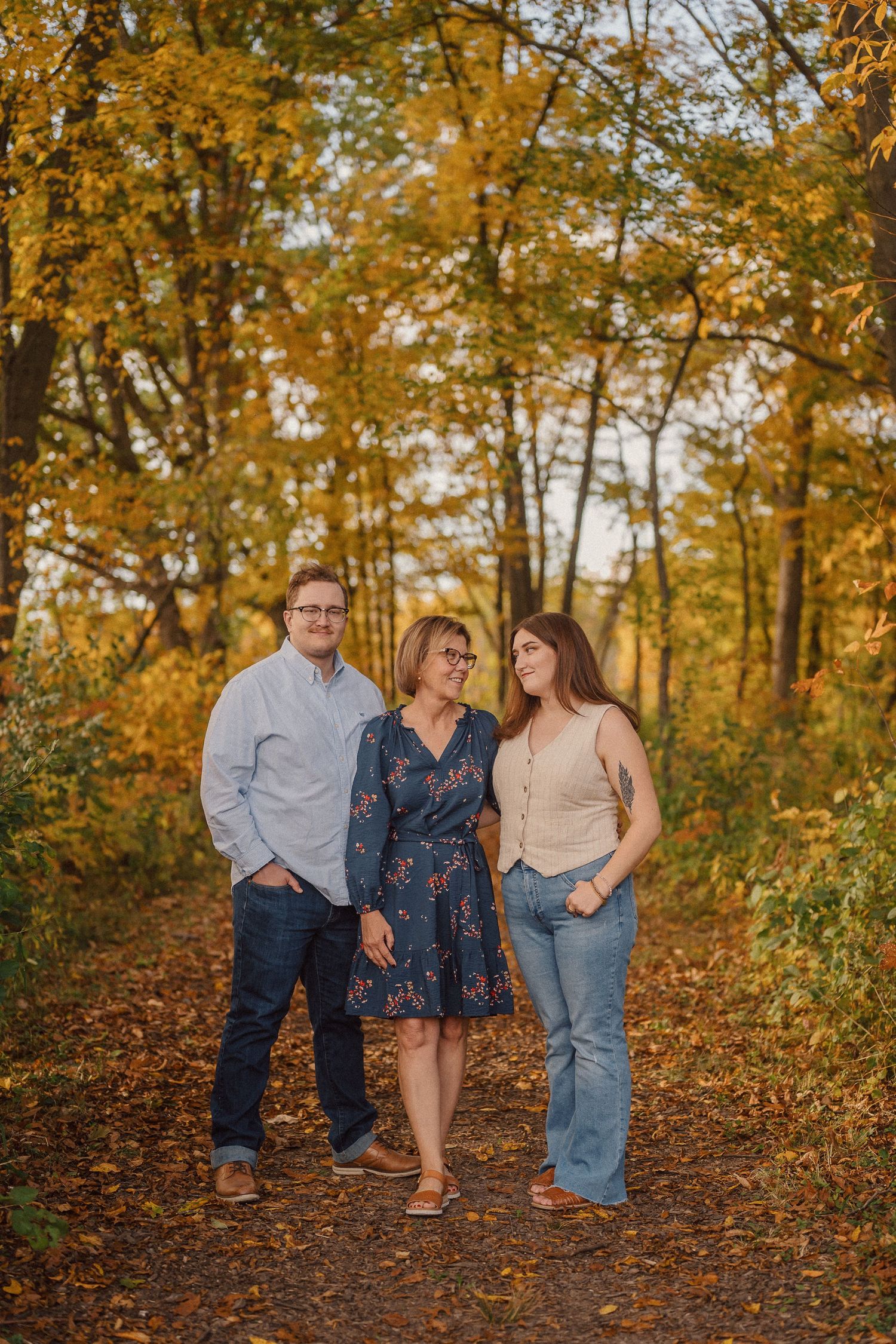 Three people stand close together on a wooded trail surrounded by golden fall foliage.