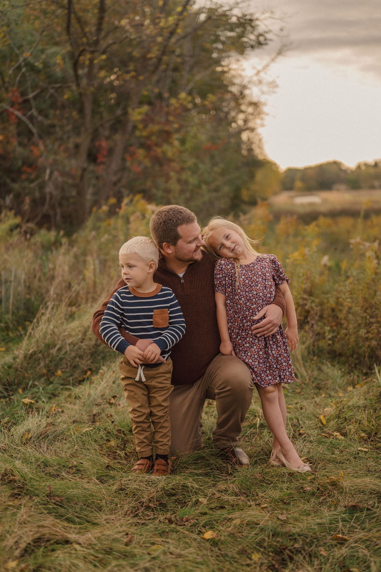 A family sits together in a grassy meadow during golden hour, surrounded by autumn foliage.