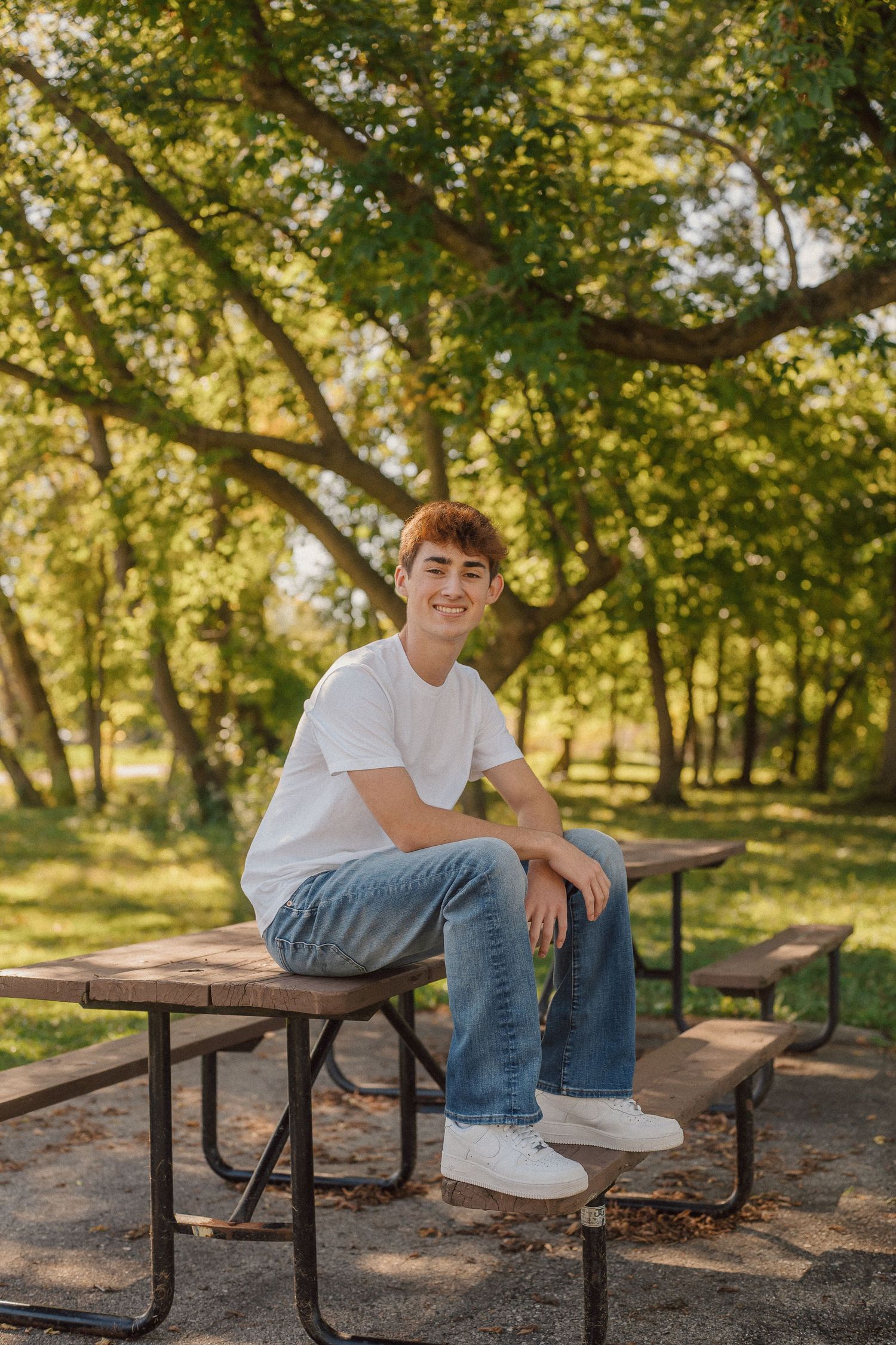 Person sitting casually on a wooden picnic table in a sunlit park with autumn trees in the background.