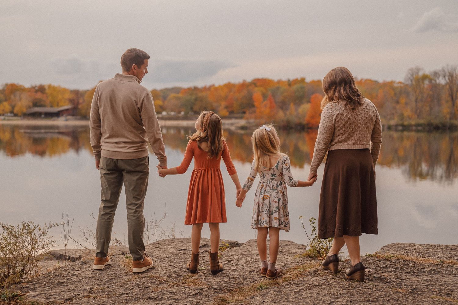 A family holds hands while looking out at an autumn lake with fall foliage in the background.