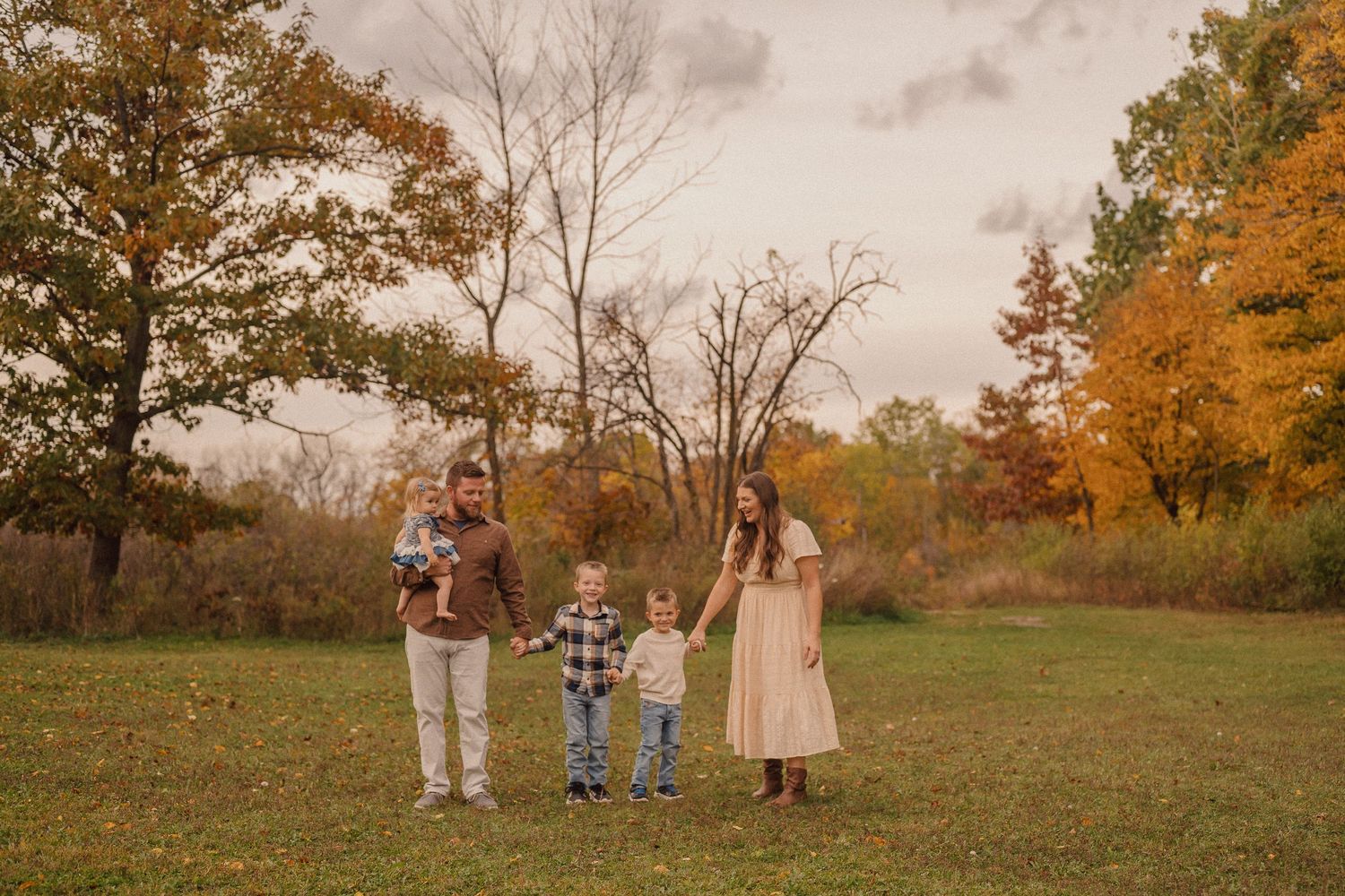 Family group standing in open field with autumn trees and golden sunset light behind them.